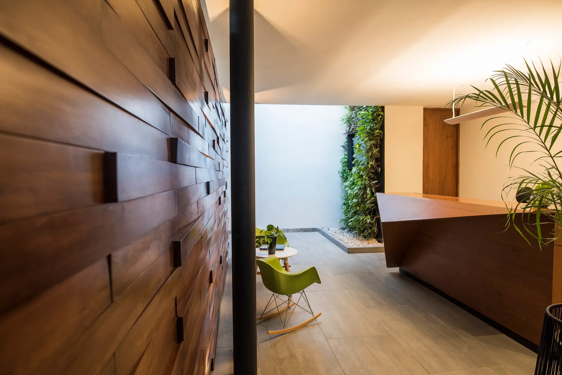 A modern reception area features a textured horizontal wood slat wall, green rocking chairs, an angular wooden desk, and a vertical living wall.