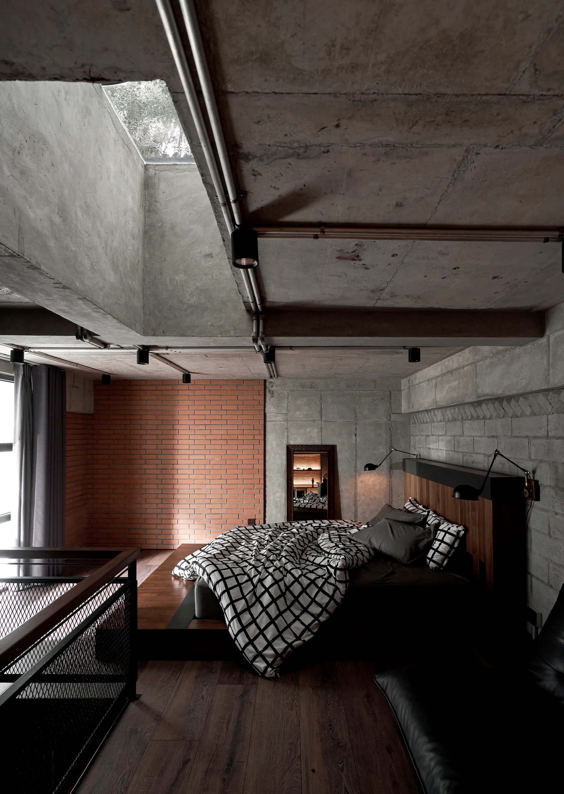 An industrial bedroom with a low wood platform bed set against raw concrete walls and a red brick accent section under a concrete ceiling with a skylight.