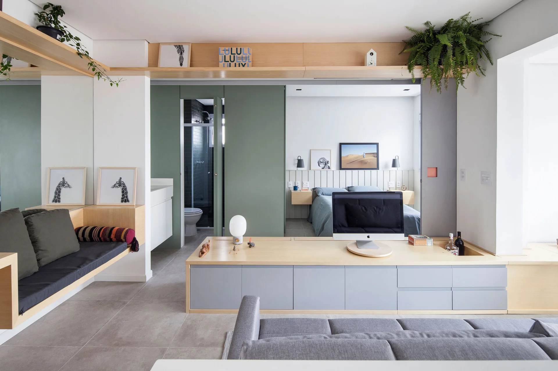 A modern bedroom view showing a light wood built-in desk, a grey sofa, and a sage green sliding door that opens to a sleeping area.
