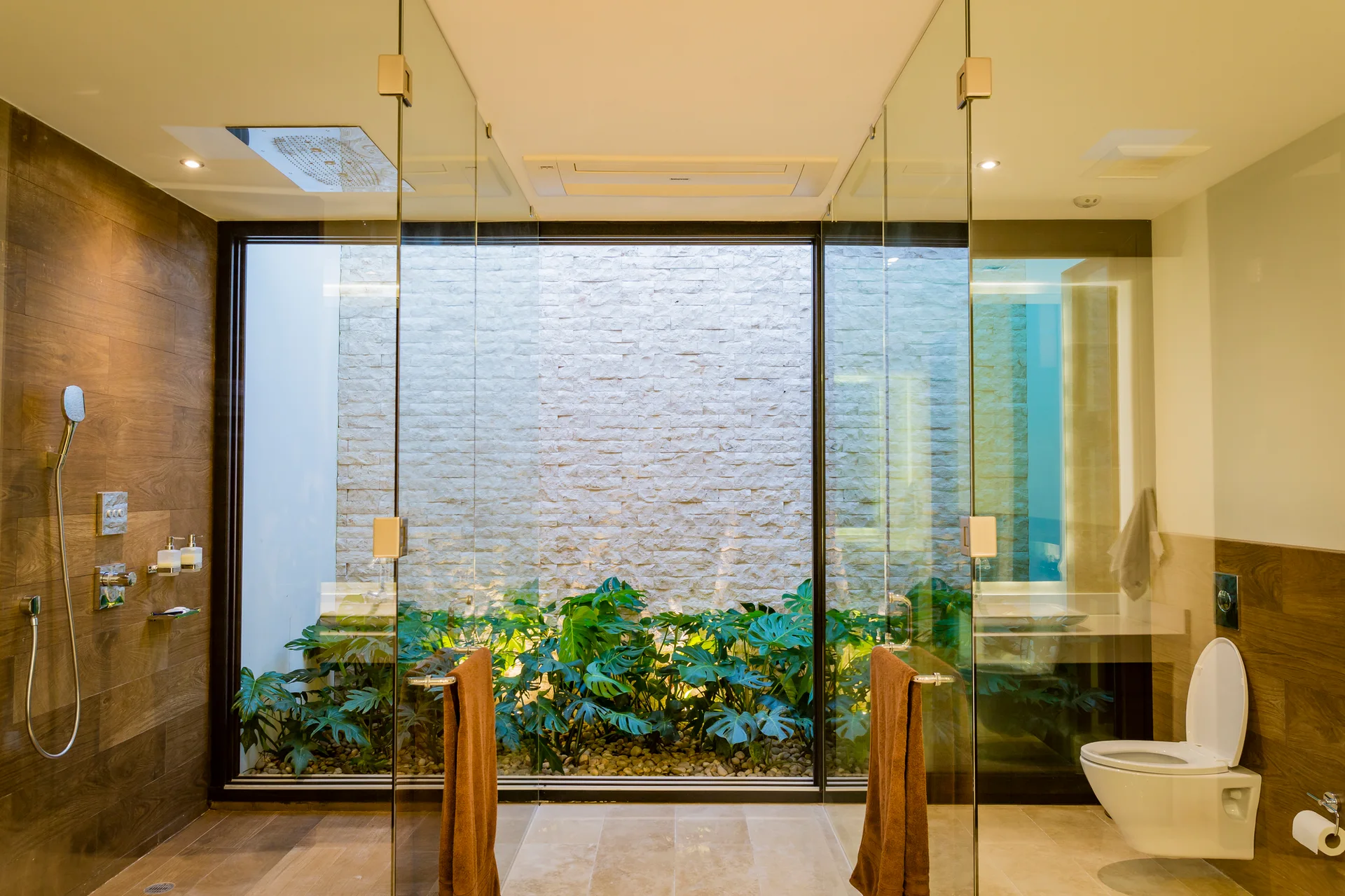 A modern bathroom features a glass-walled shower looking out onto a private atrium garden with leafy monstera plants and a stacked stone wall.