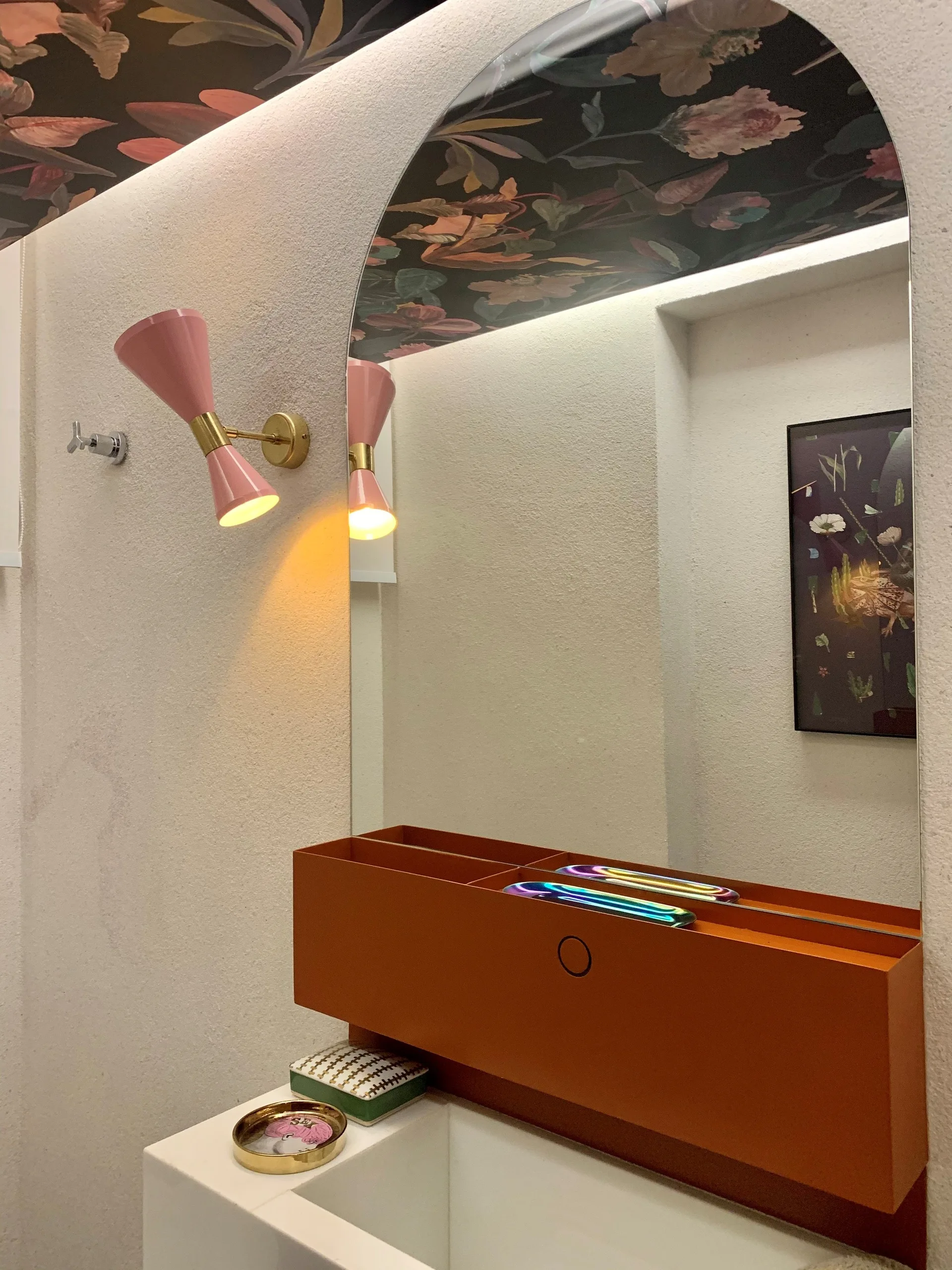 A white stucco powder room featuring a large arched mirror, a bright orange vanity shelf, a pink wall sconce, and dark floral ceiling wallpaper.