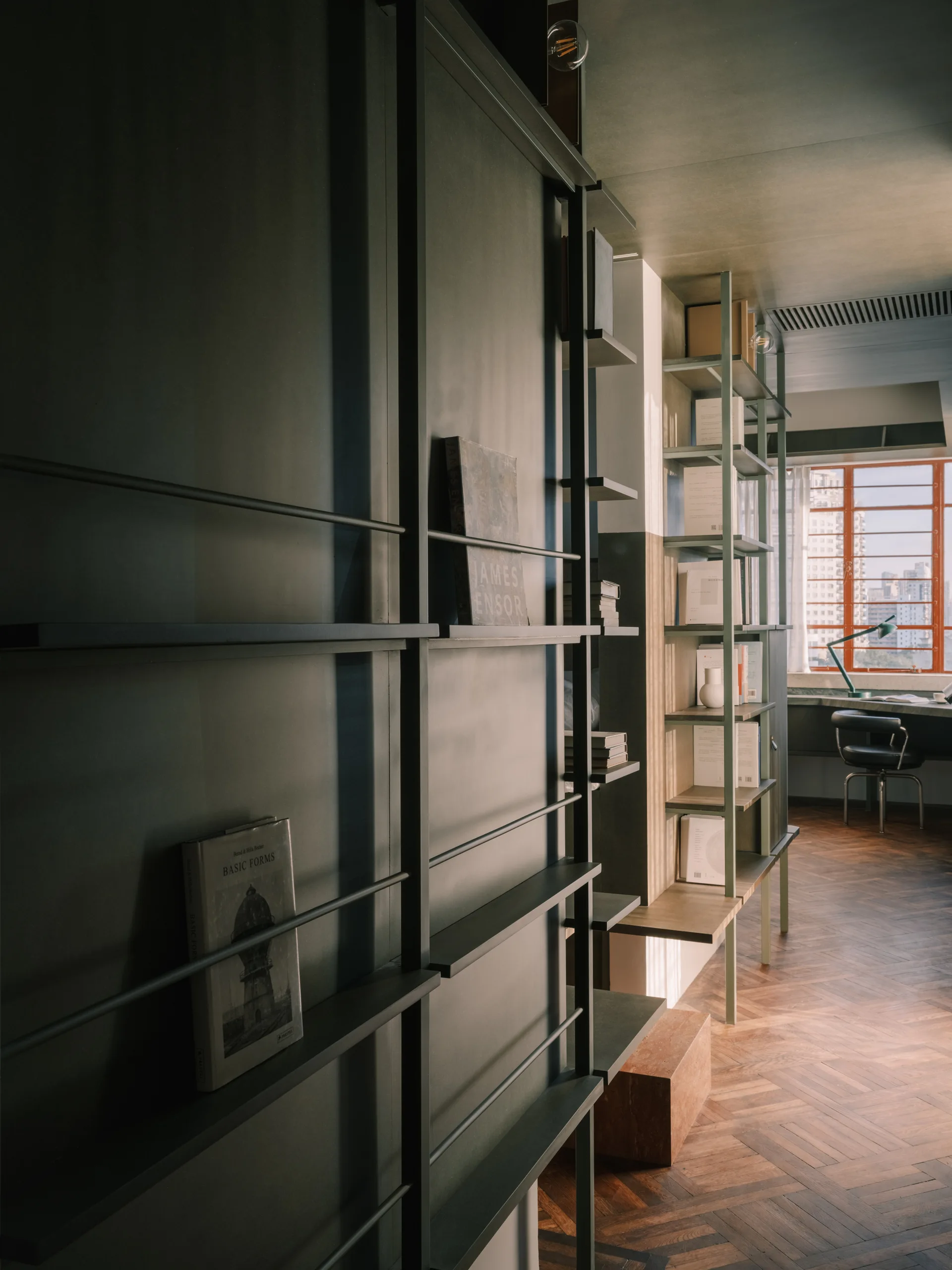 A dark green home office features tall metal built-in shelving, herringbone patterned wood floors, and a desk positioned near a grid window.