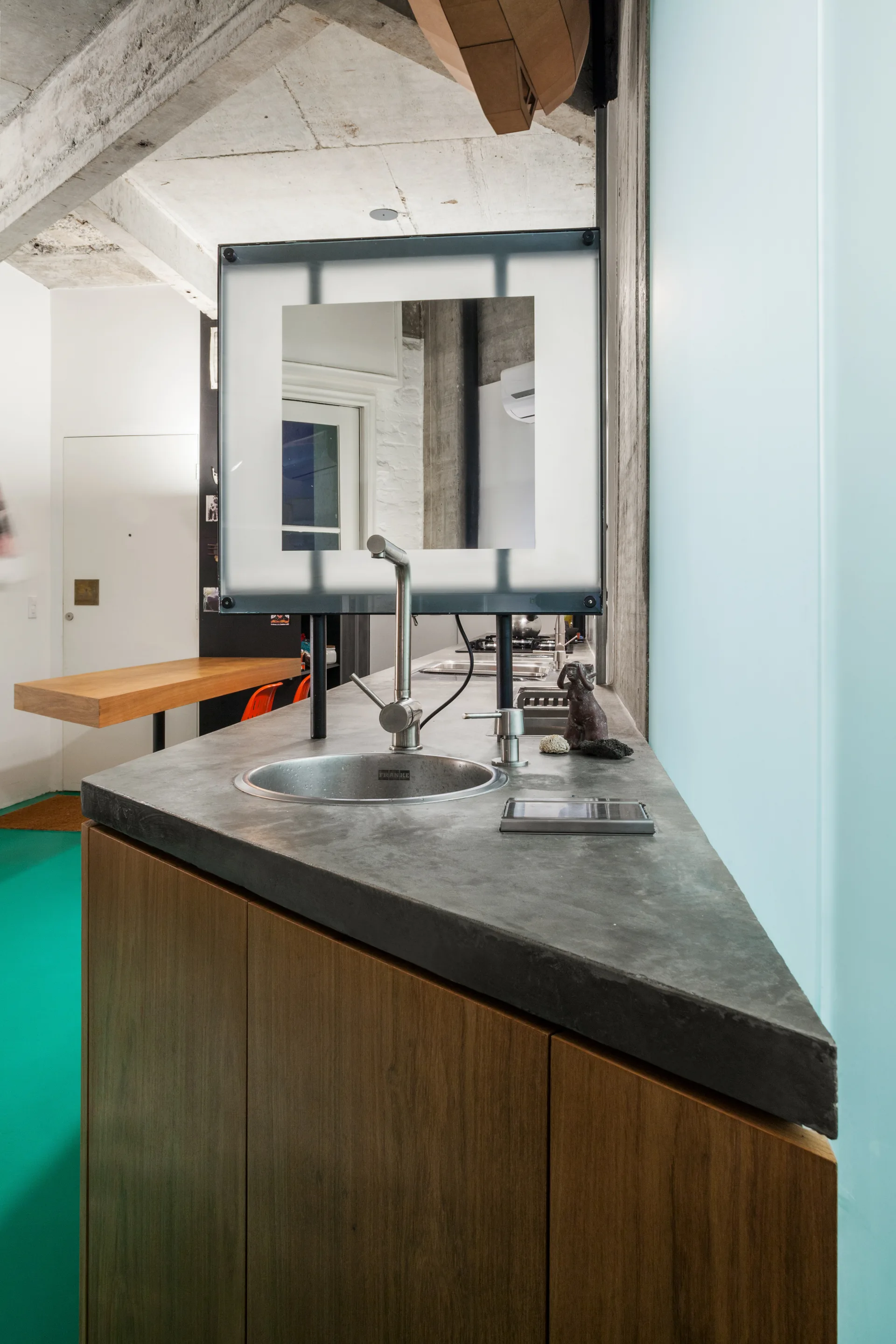 An angled kitchen island features a smooth grey concrete countertop with an integrated circular sink and a mounted square mirror above it.