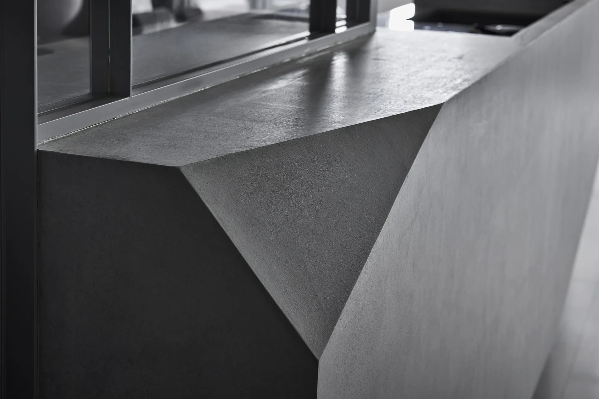 A close-up view of a custom gray concrete built-in with sharp geometric angles situated beneath a dark metal-framed glass partition.