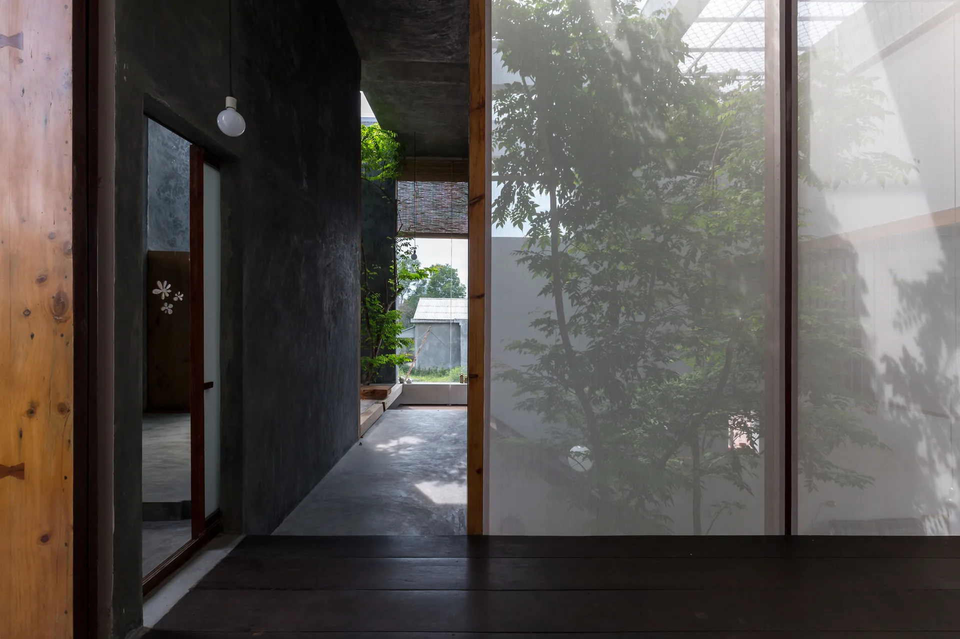 A dark concrete hallway with a wooden doorway features a tall translucent screen showing the silhouette of an indoor courtyard tree.