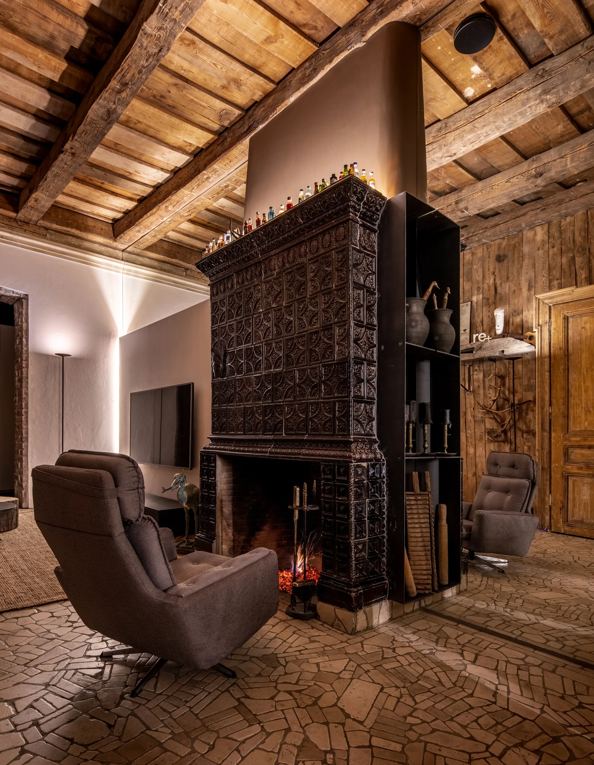 A rustic living room featuring a large dark tiled fireplace centerpiece flanked by gray armchairs under an exposed wood beam ceiling.