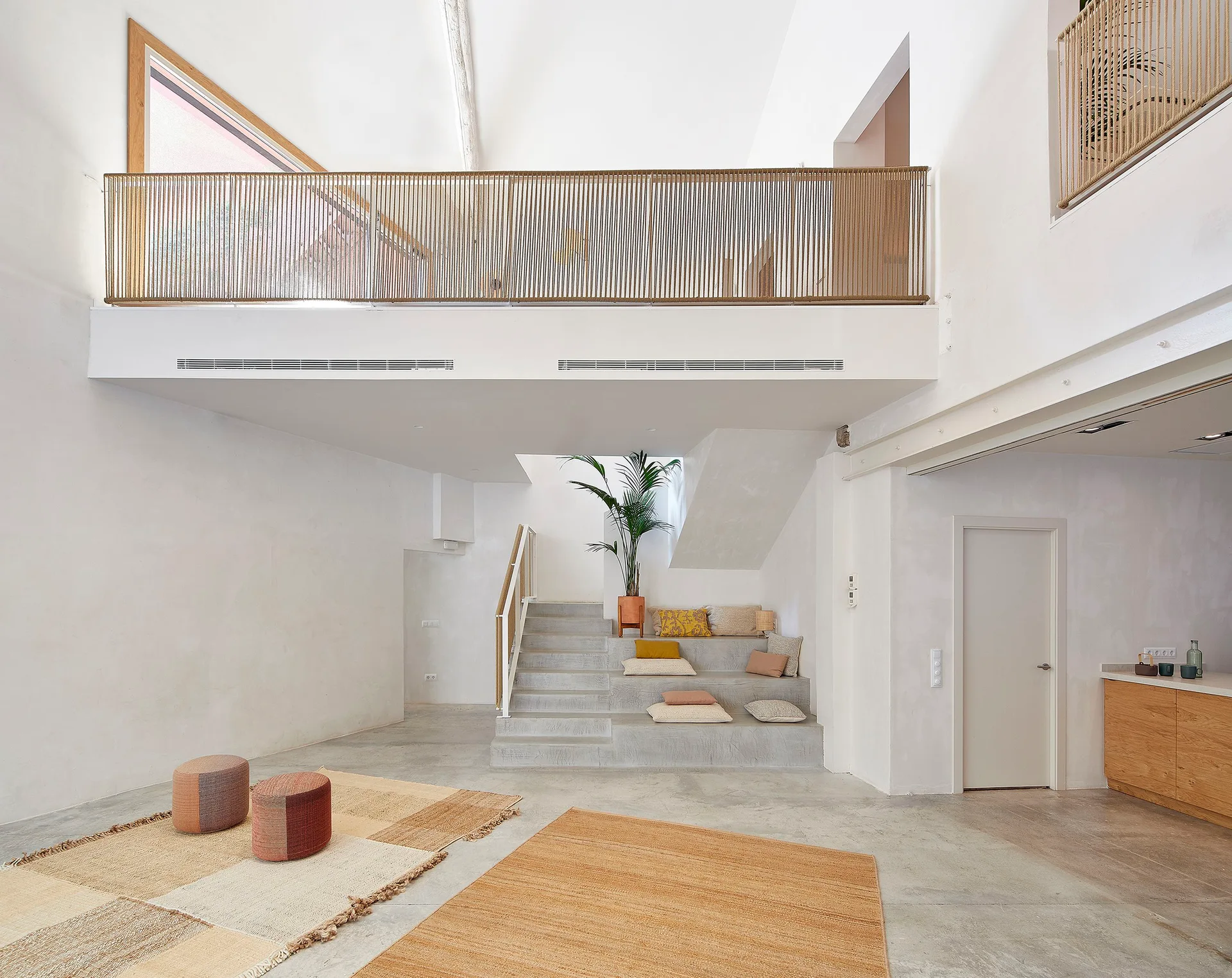 A minimalist interior features a wide concrete staircase with integrated stepped seating, woven floor rugs, and a mezzanine balcony with a rope railing.