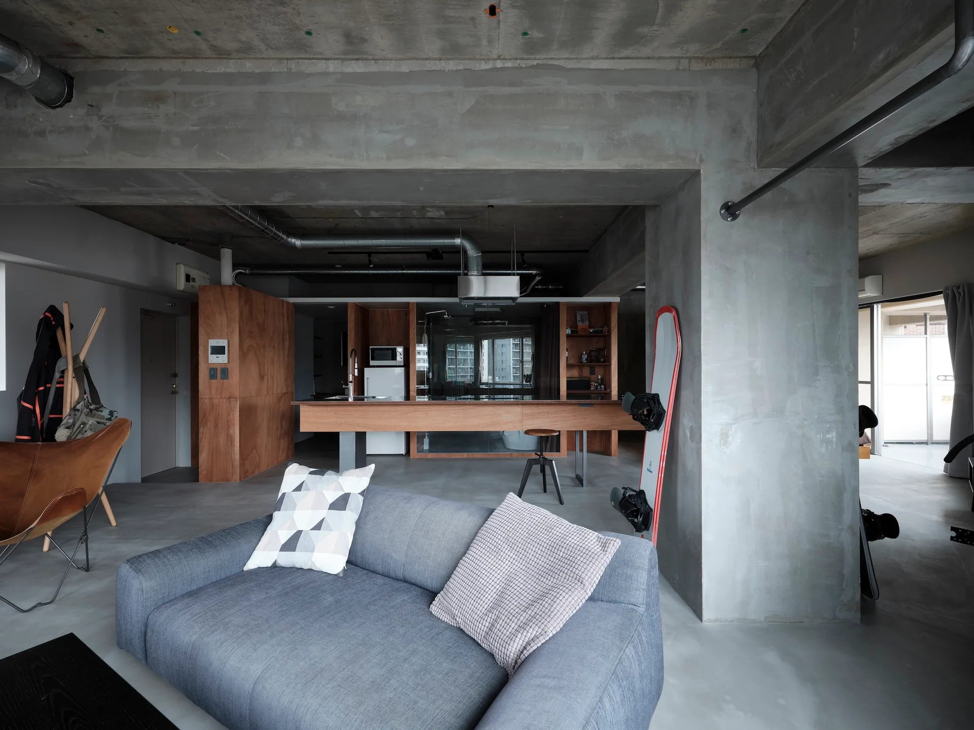 An industrial style open apartment features a grey fabric sofa, raw concrete walls and ceiling, and a wooden kitchen island.