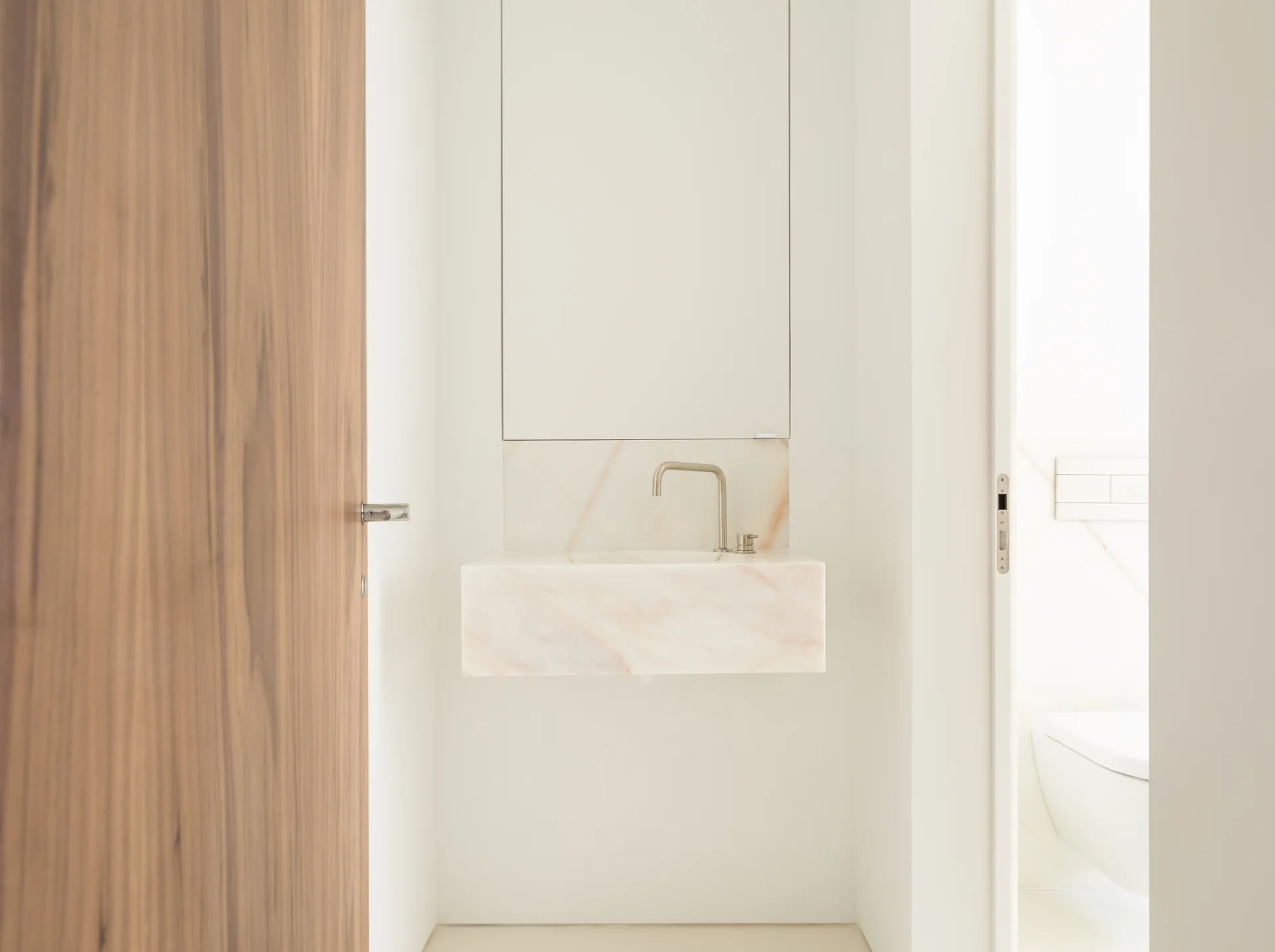 A minimalist powder room with a wall-mounted veined stone block sink, a silver faucet, and a warm wood door.
