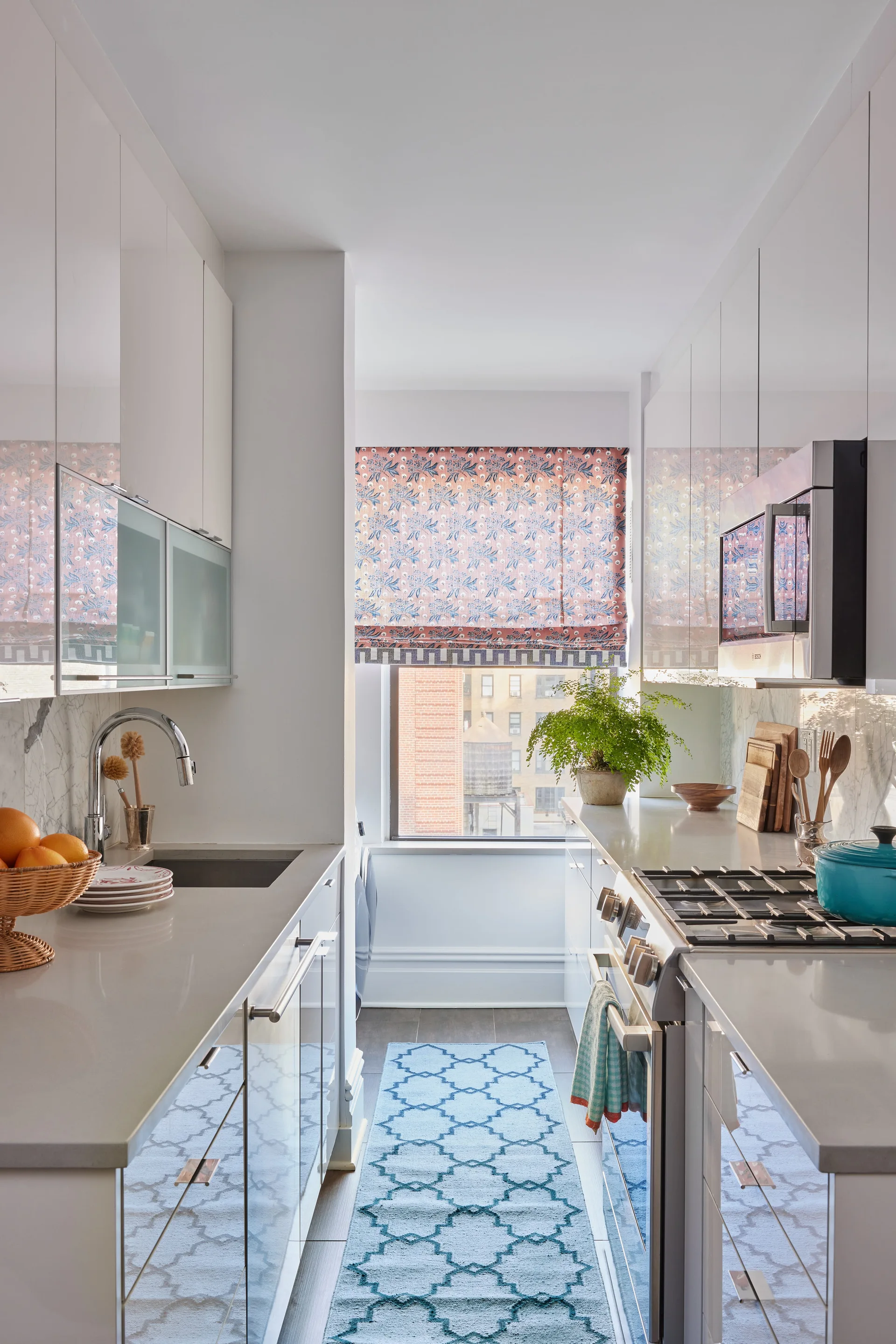 A narrow galley kitchen features glossy white cabinetry, stainless steel appliances, a blue patterned runner rug, and a floral Roman shade on the window.