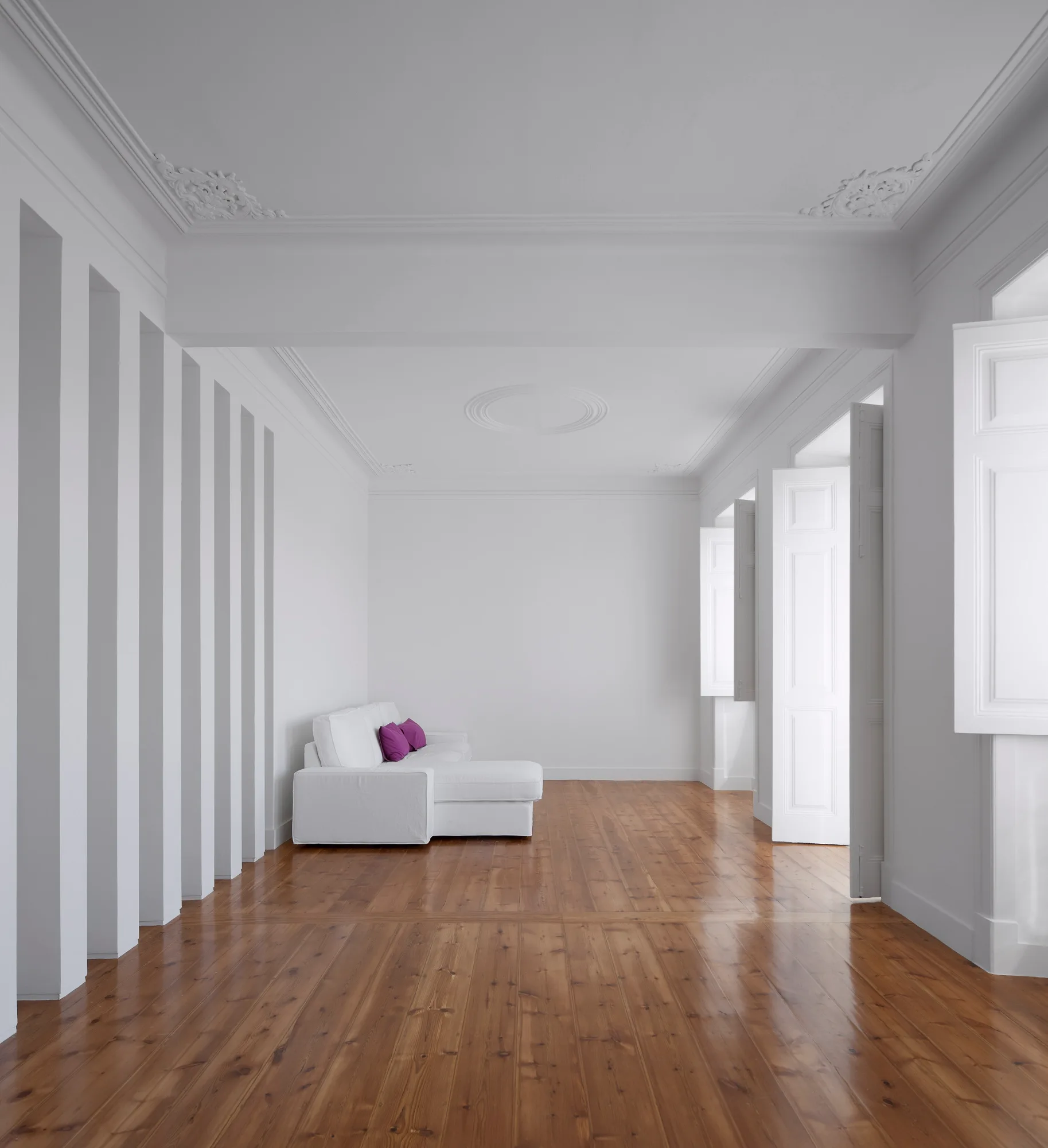A spacious white room features vertical wall paneling, a ceiling medallion, polished wood flooring, and a single white sofa with purple pillows.