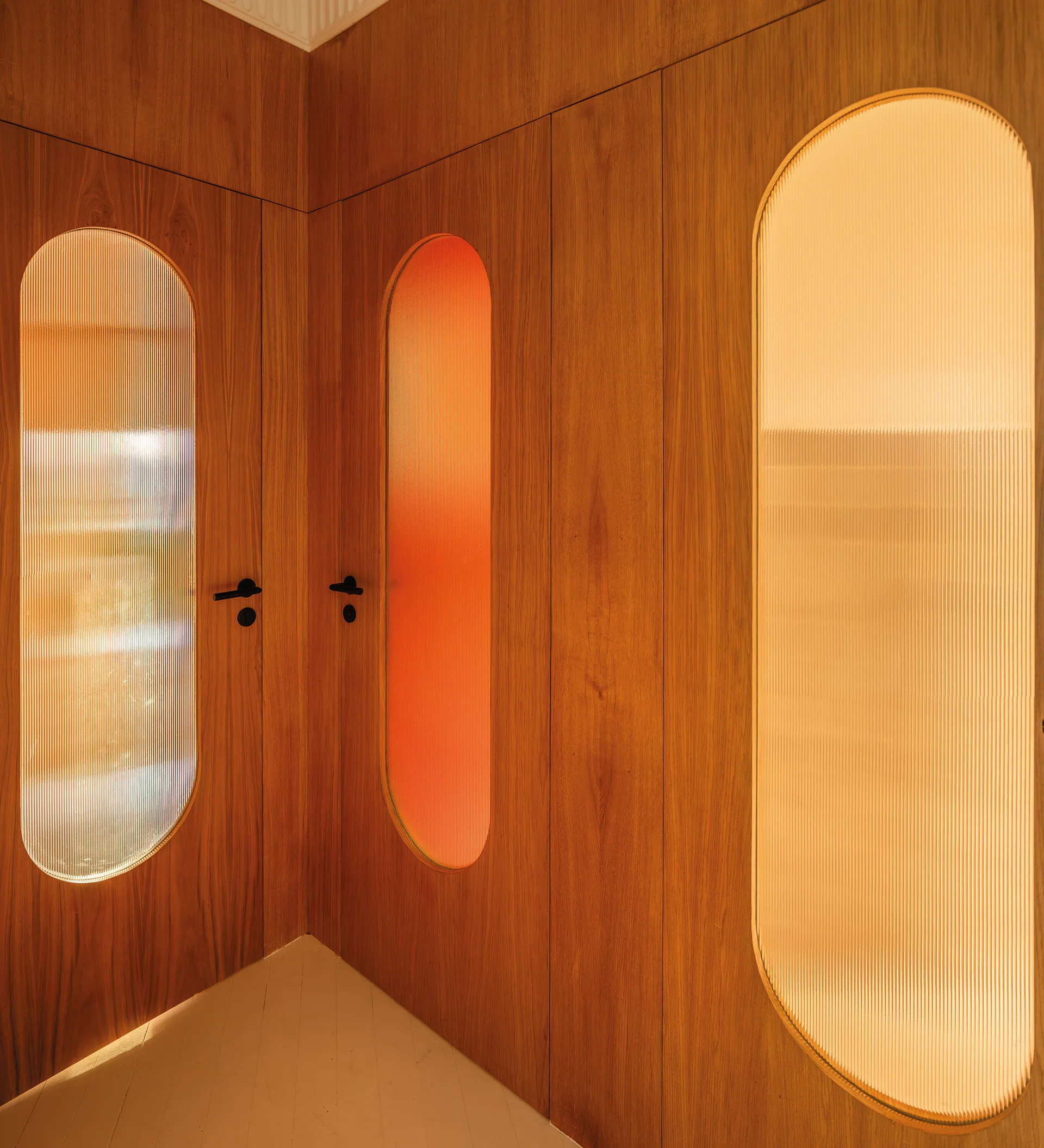 A hallway featuring flush wooden doors with tall, arched fluted glass panels backlit with warm orange and yellow light.