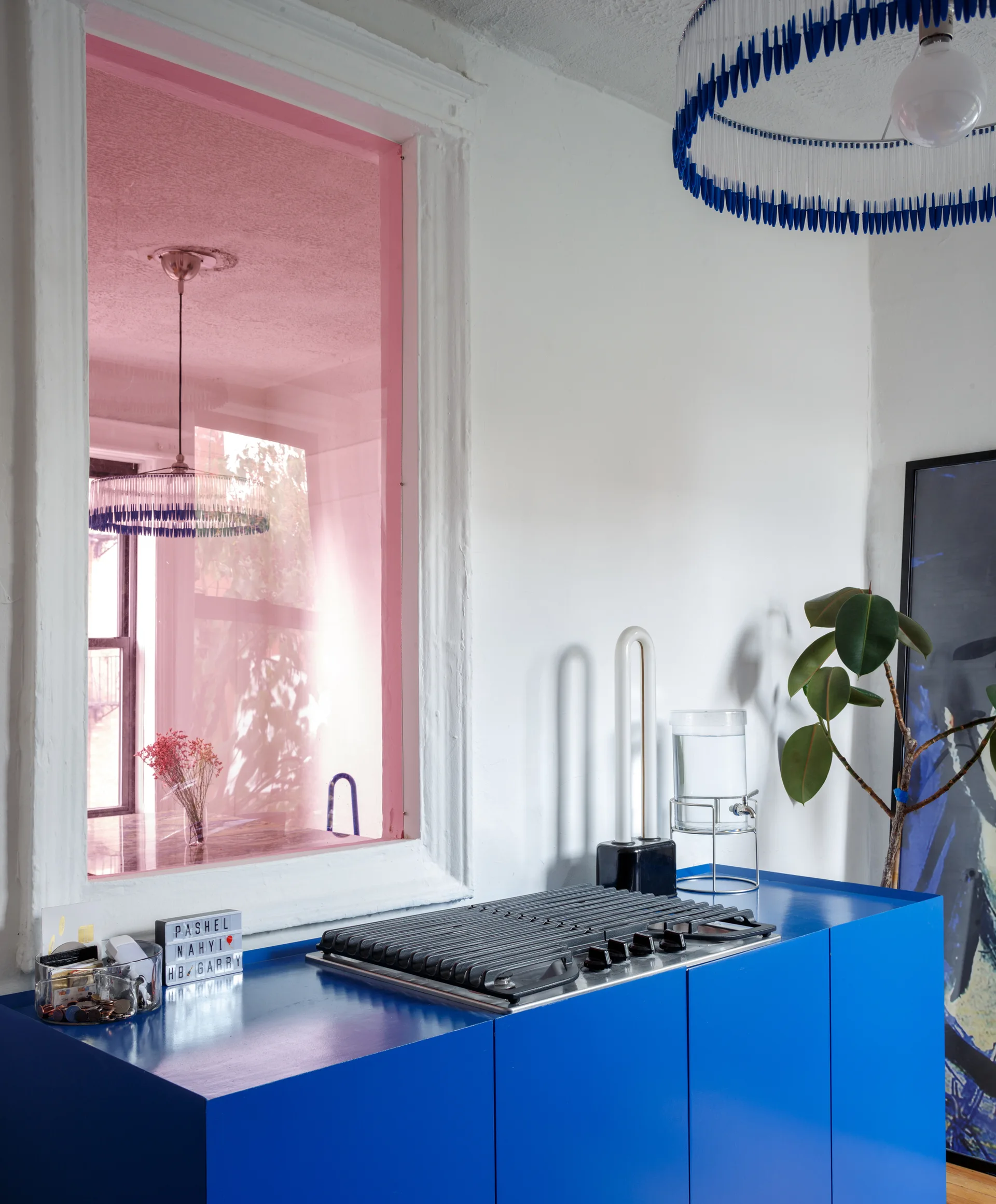 A bright blue kitchen cabinet features a built-in black stovetop beneath a white wall with a pink-tinted window looking into another room.