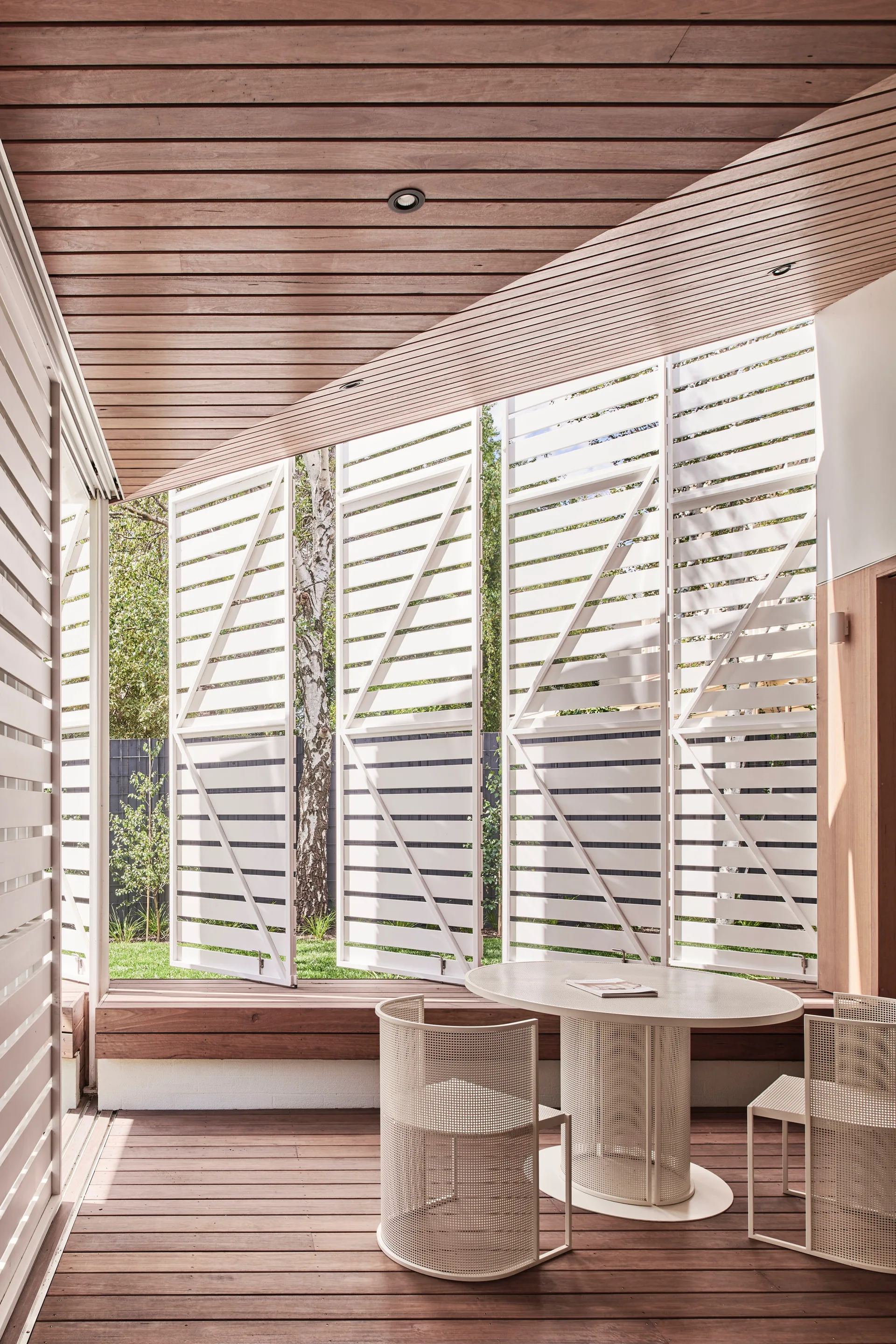 A covered wood patio features a white mesh dining table and chairs set against white slatted privacy shutters opening to a garden.