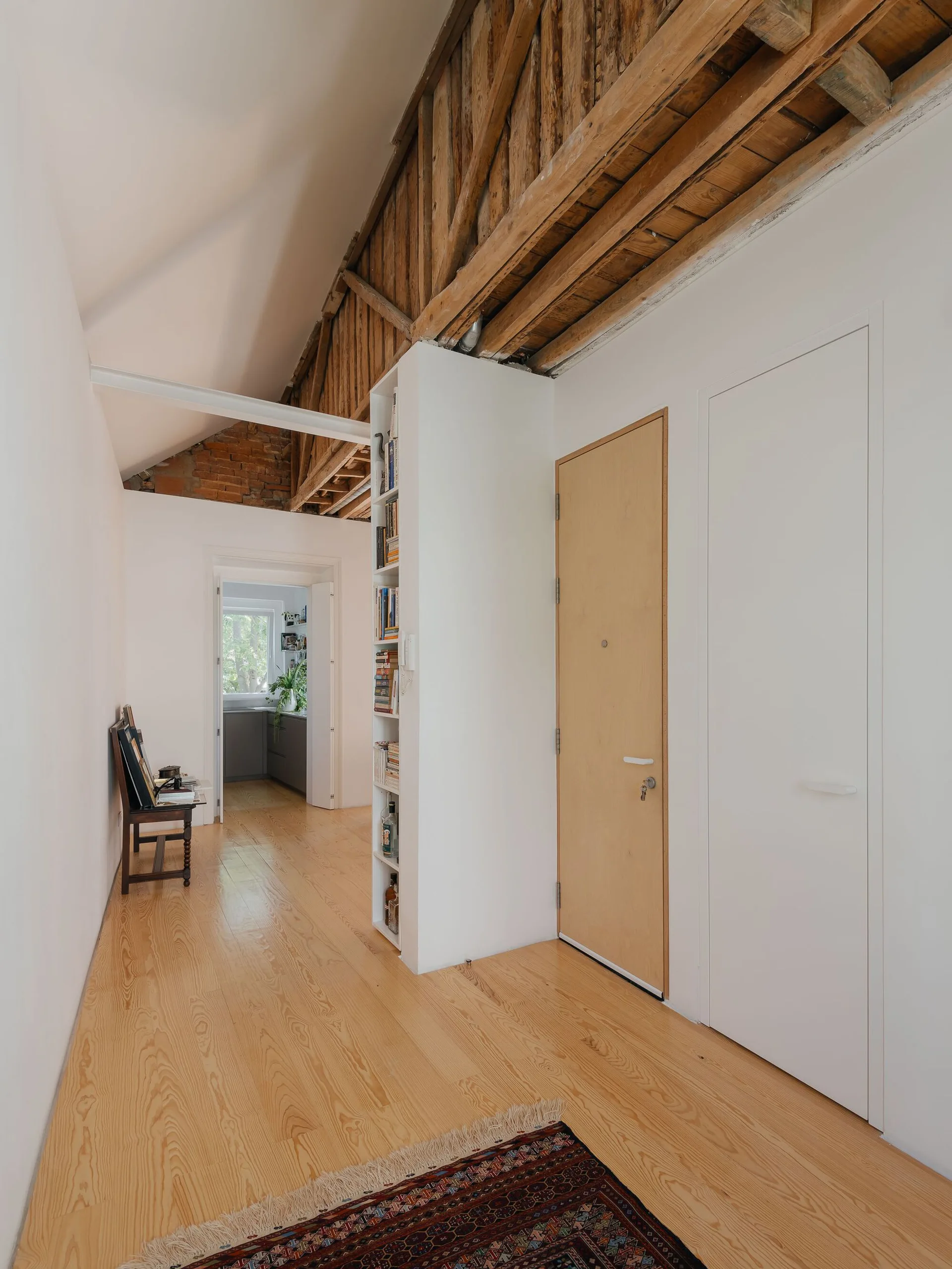 A bright hallway featuring light wood floors, raw wooden ceiling trusses, smooth white walls, and a built-in vertical bookshelf.