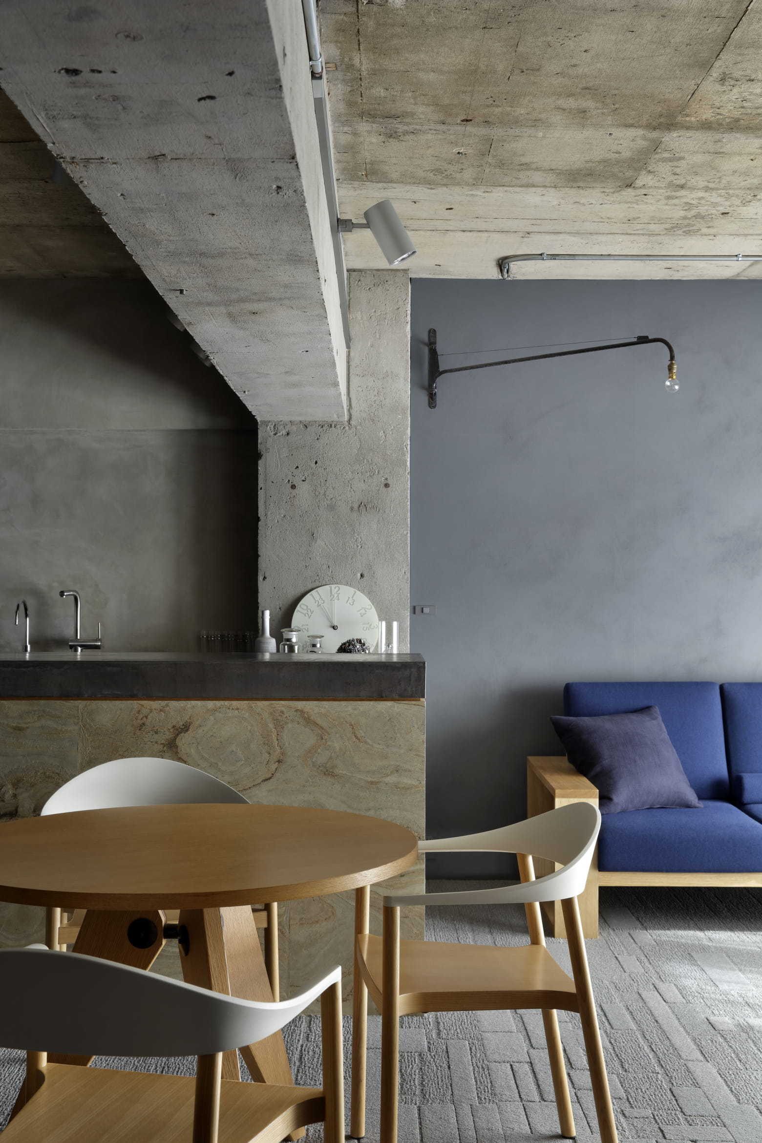 An industrial living area features an exposed concrete ceiling and beam, a blue fabric sofa, and a round wooden dining table with chairs.
