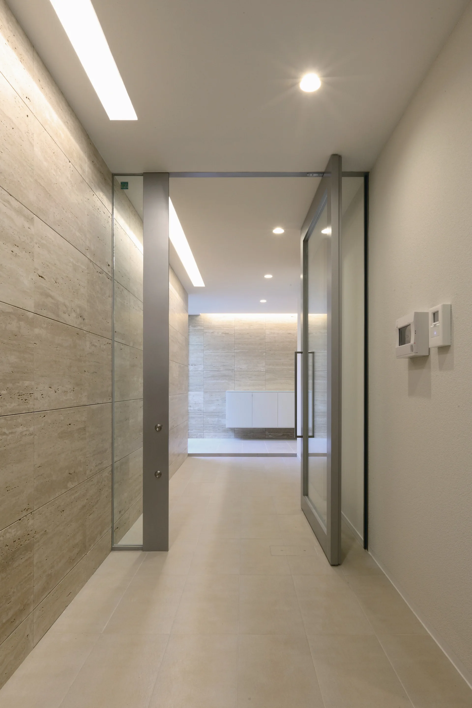 A minimalist hallway features a beige stone tiled wall, a glass entry door in a grey frame, and recessed ceiling lighting.