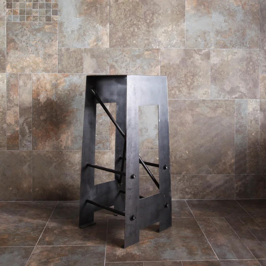 A dark raw metal bar stool with cross-bracing sits on a matching stone-look tile floor in front of a slate-like tiled wall.