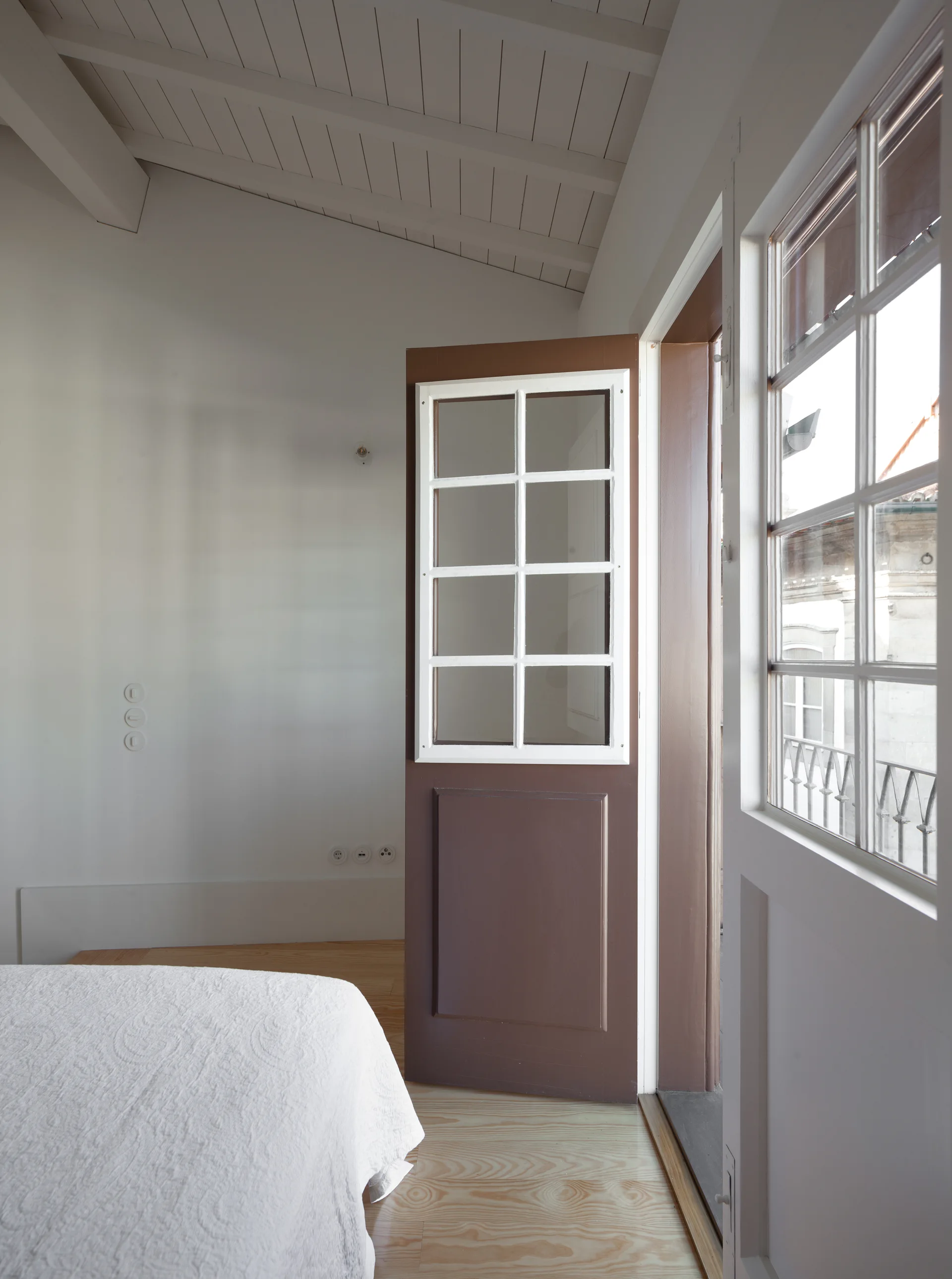 A minimalist bedroom featuring white walls, a sloped wood ceiling, a bed with a white quilt, and an open brown door with a glass pane grid.