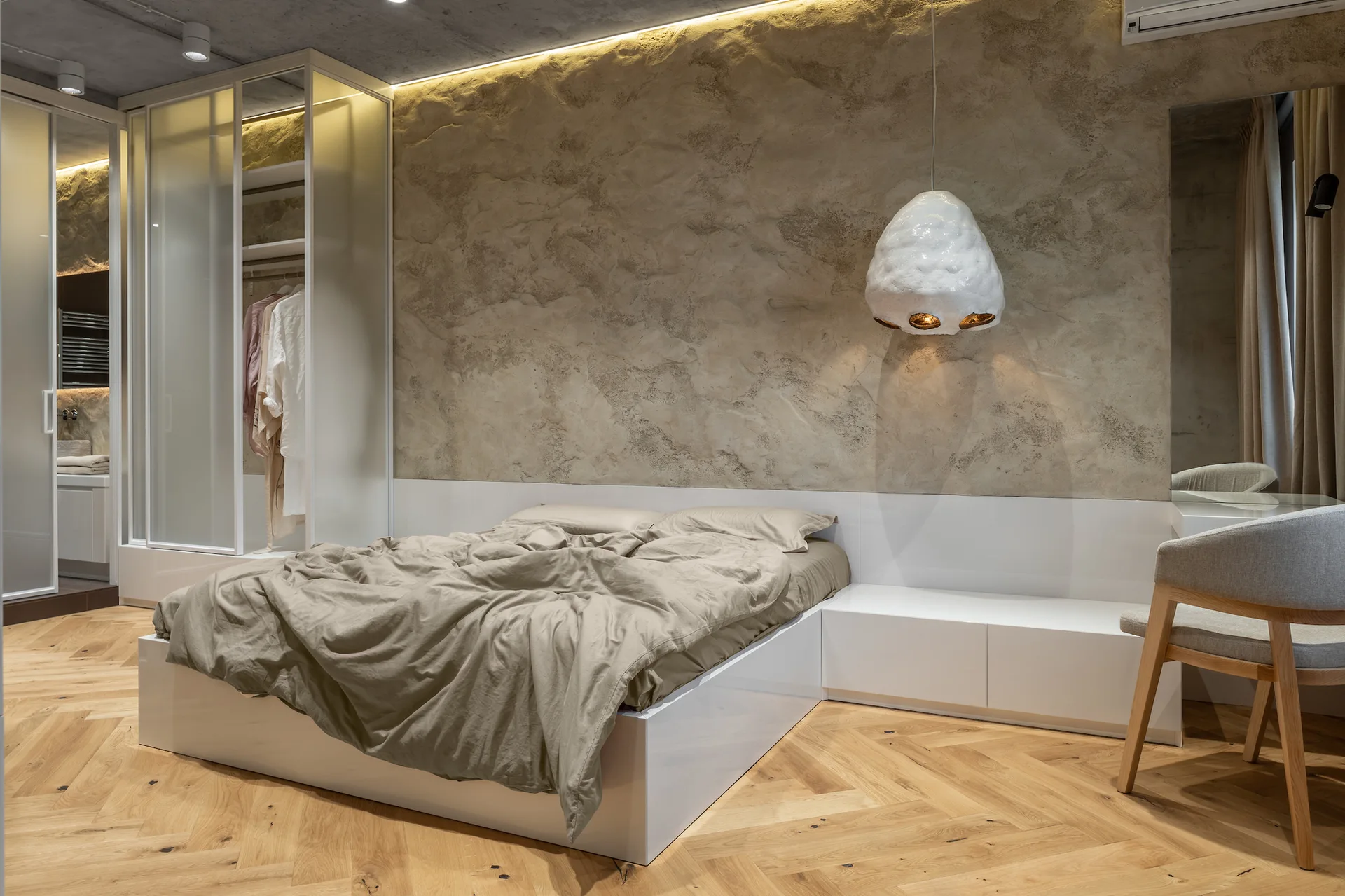 A modern bedroom features a low white platform bed against a textured beige plaster wall, herringbone wood floors, and a translucent glass wardrobe.