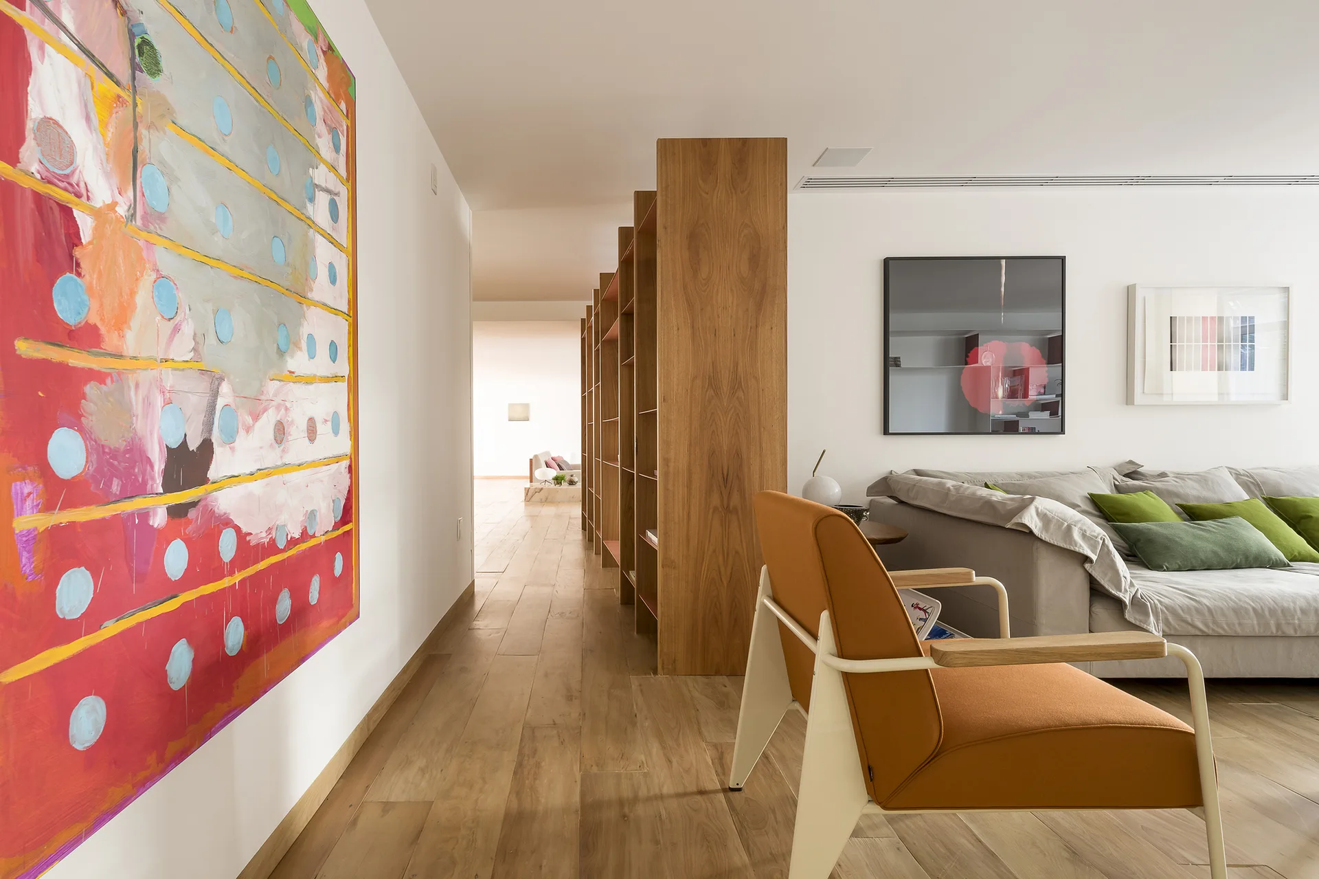 A modern living room featuring a tall wood shelving unit used as a room divider, a grey sofa with green pillows, an orange armchair, and abstract wall art.