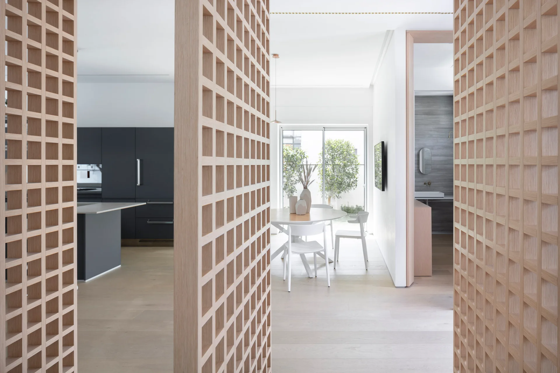 Light wood lattice room dividers frame a minimalist dining area with a round white table and a dark grey kitchen in the background.