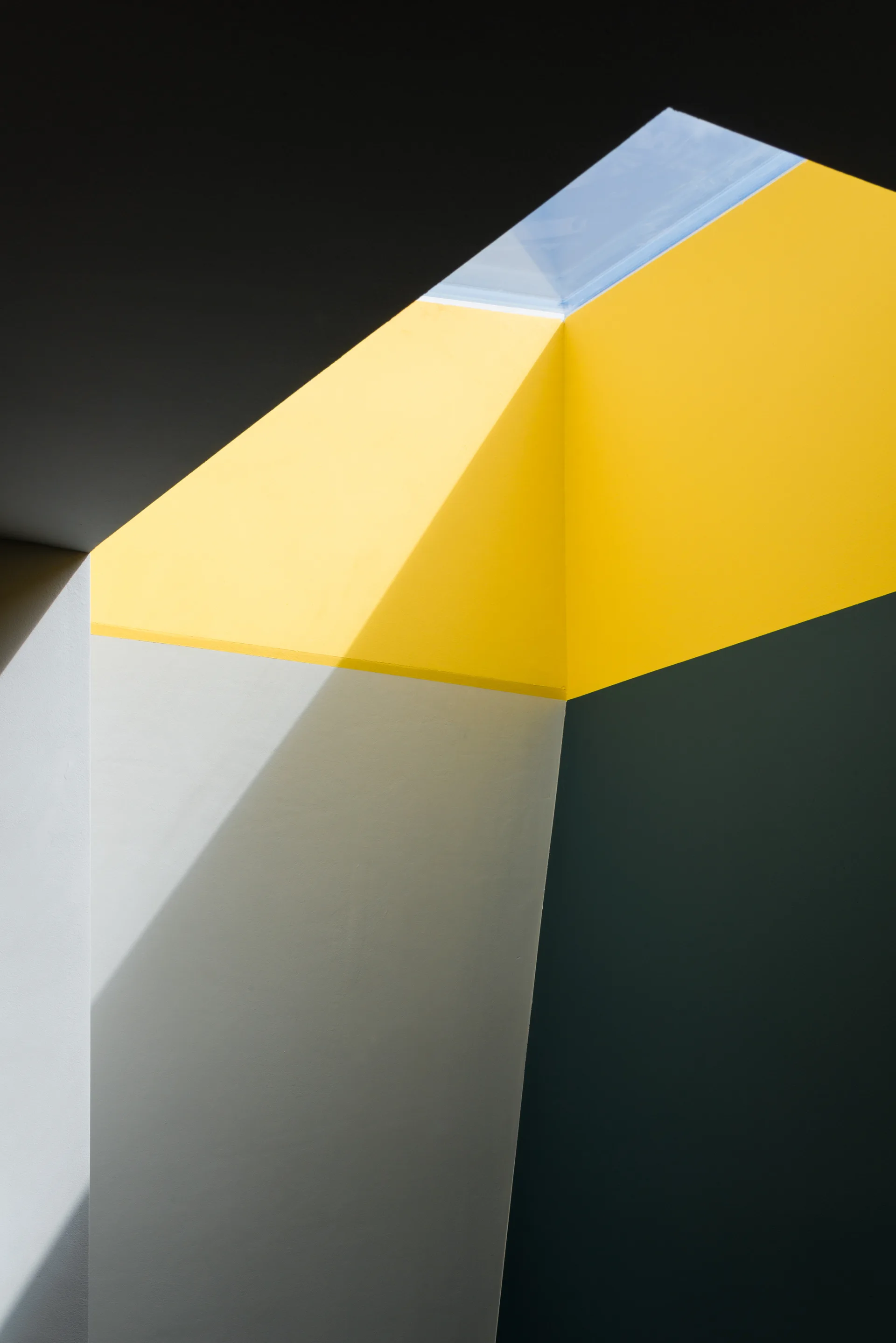 An architectural light well features intersecting angled walls painted in matte yellow, grey, and dark green beneath a small skylight.