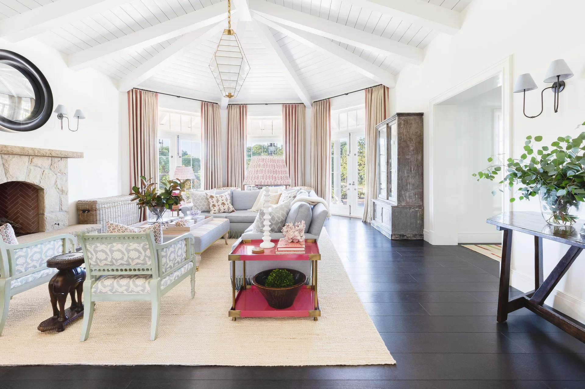 A bright living room features a white vaulted beamed ceiling, a light blue sectional sofa, pale green armchairs, and a stone fireplace.