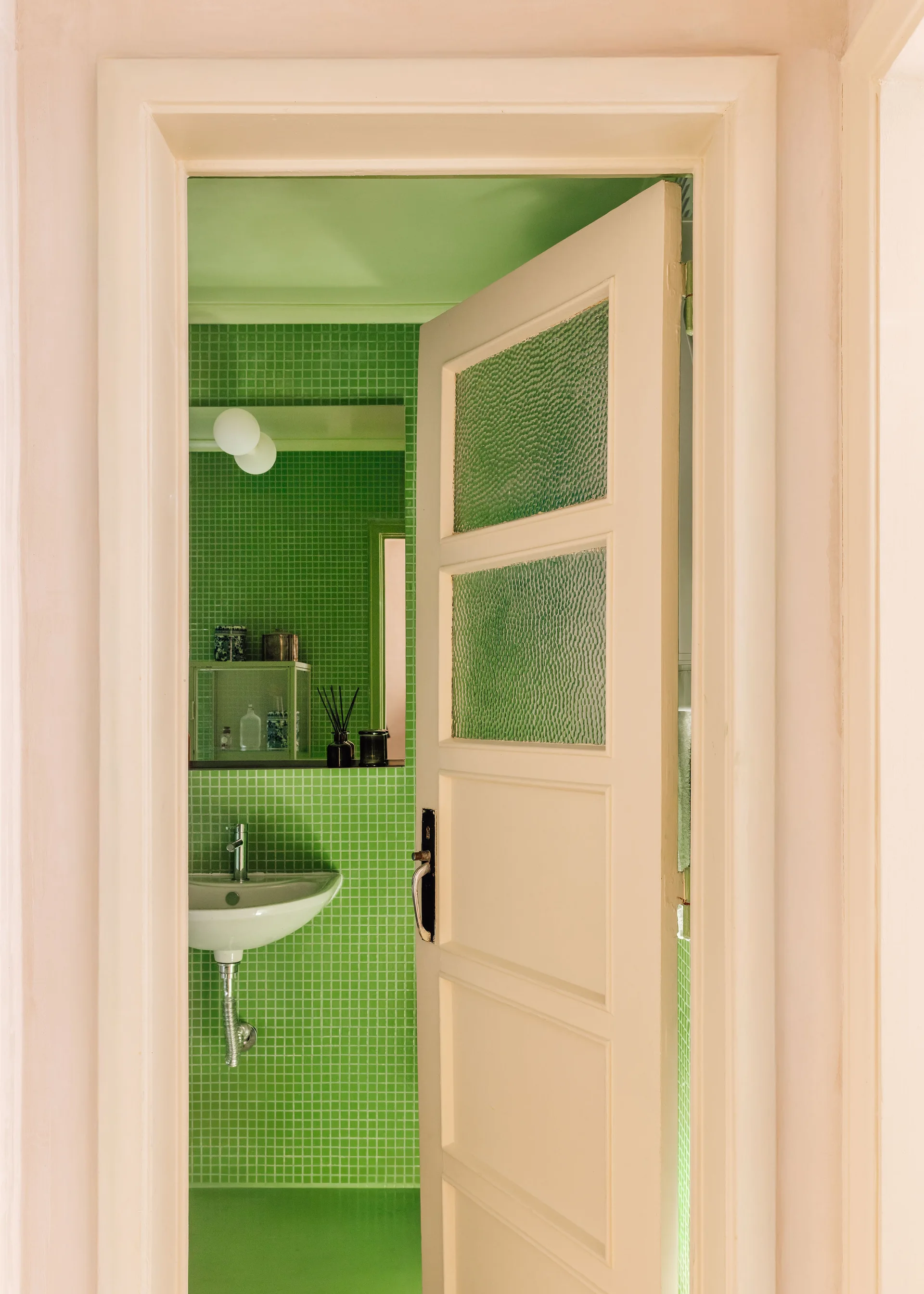 An open cream-colored door with textured glass panels reveals a small bathroom with bright green mosaic tile walls and a white sink.