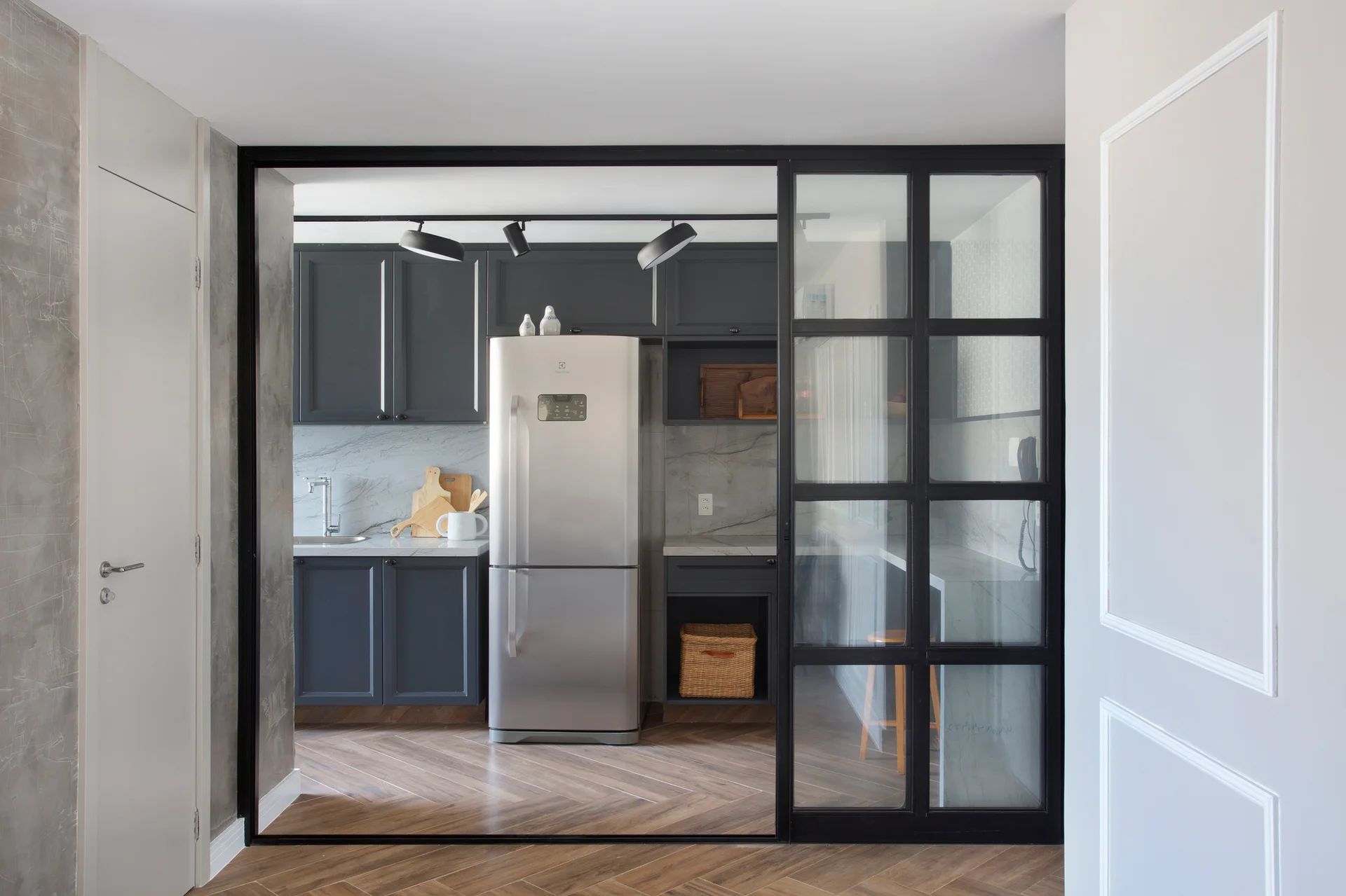A kitchen with dark grey cabinets and a stainless steel refrigerator is viewed through a black-framed glass sliding partition over herringbone wood floors.