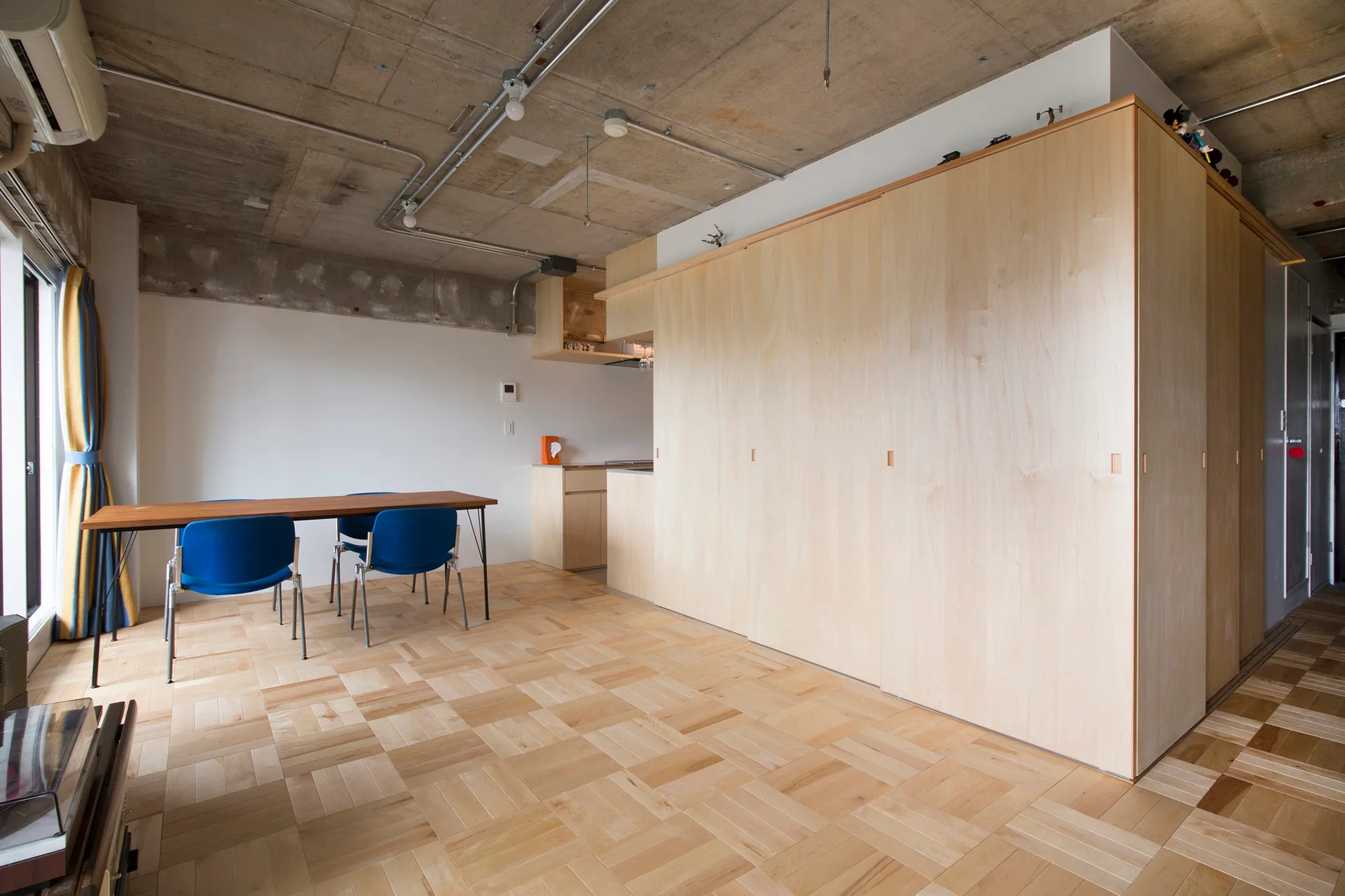 A dining room features a large light wood sliding-door storage cabinet, parquet flooring, a wood table with blue chairs, and an exposed concrete ceiling.