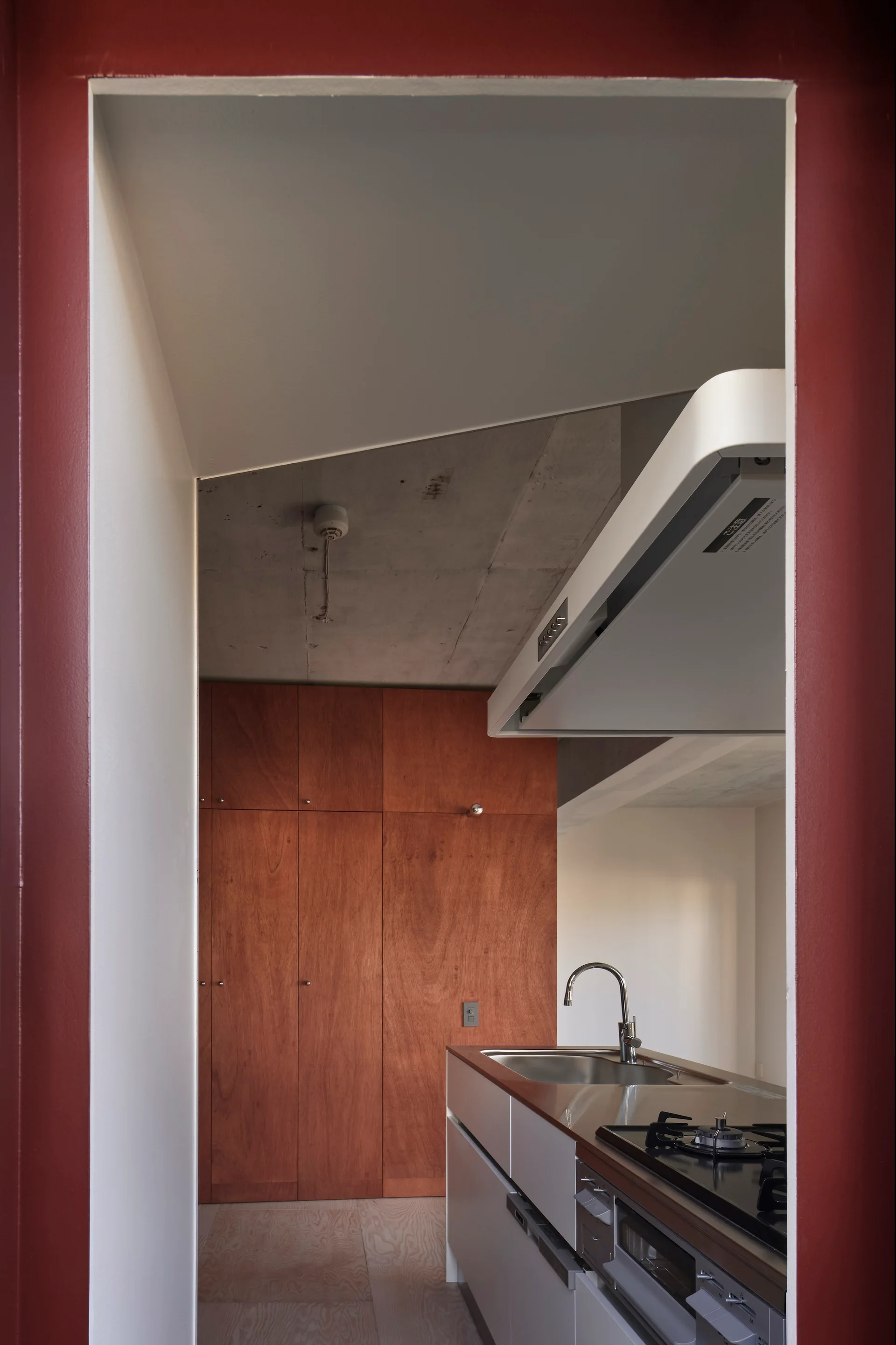 A view through a deep red doorway into a modern kitchen with a stainless steel countertop, wood cabinetry, and an exposed concrete ceiling.