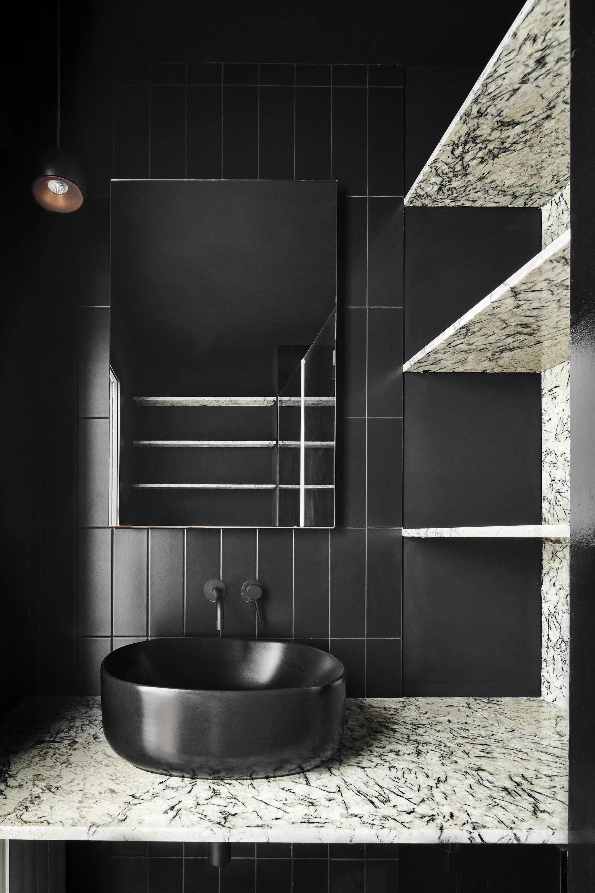 A black vessel sink sits on a heavily veined stone countertop next to matching stone floating shelves against a black tile wall.