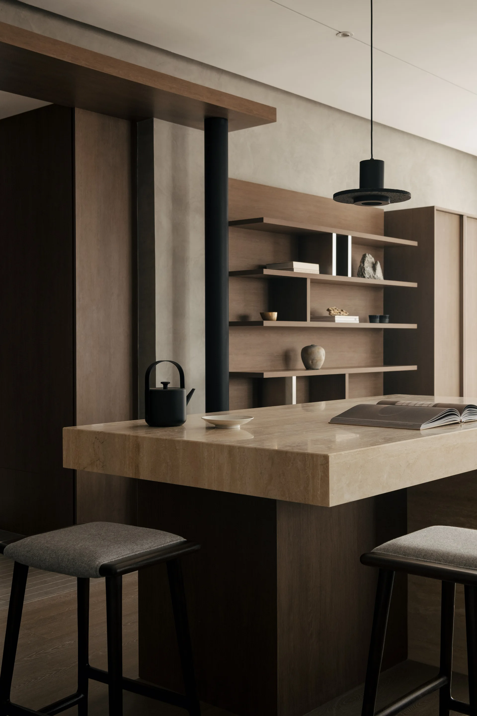 A thick stone kitchen island with grey barstools sits in front of built-in wooden shelving under a minimalist black pendant light.