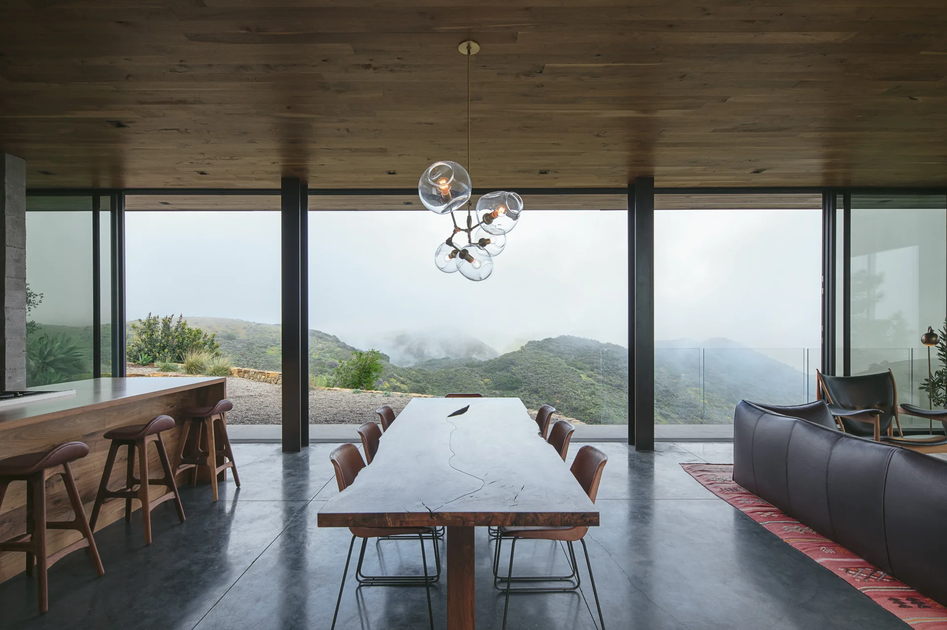 A modern dining room features a live edge wood table, leather chairs, and a wood ceiling, looking out through large windows to foggy green hills.