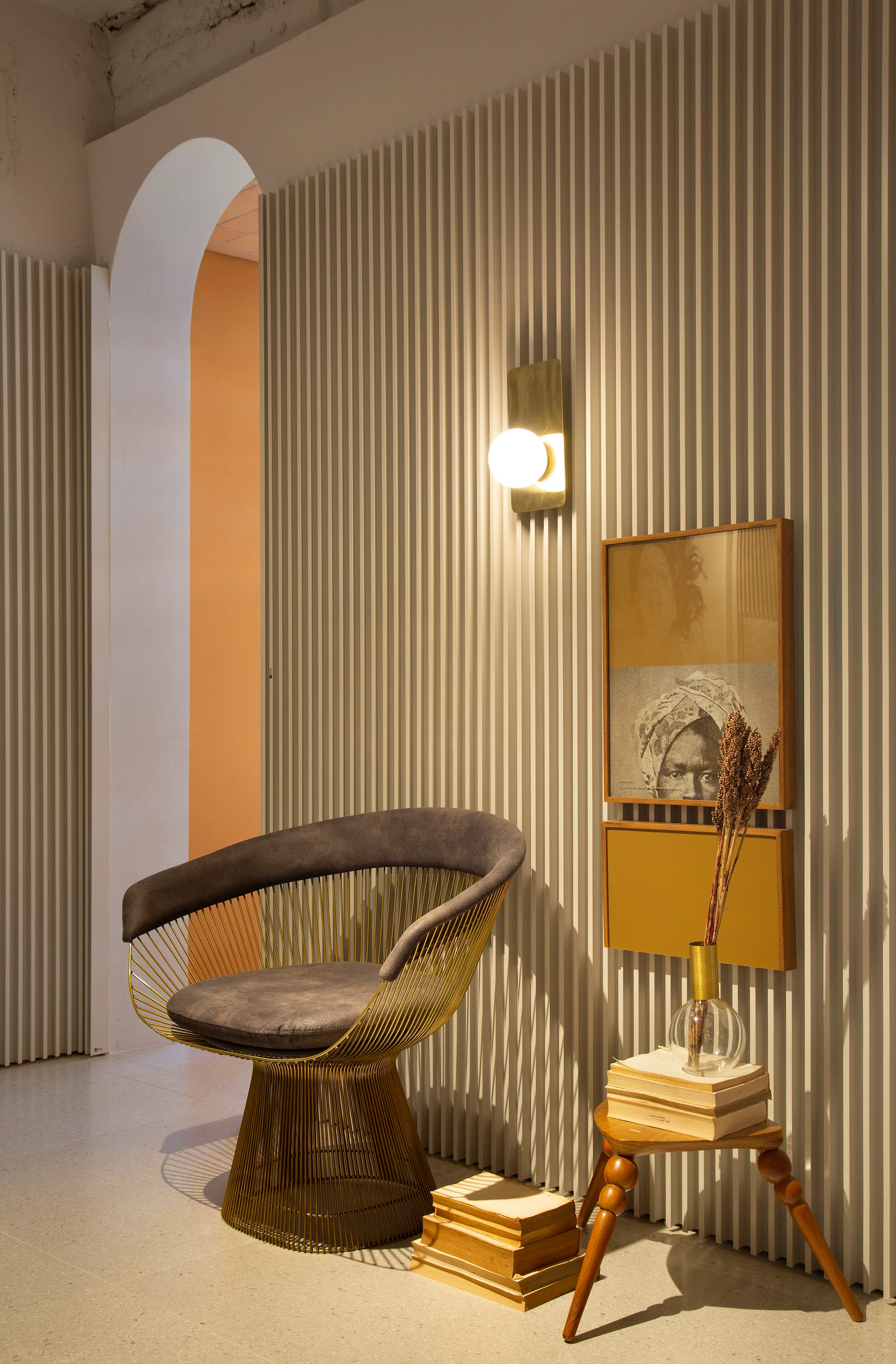 A modern gold wire accent chair sits in front of a beige fluted wood wall panel beside a glowing globe sconce and stacked vintage books.