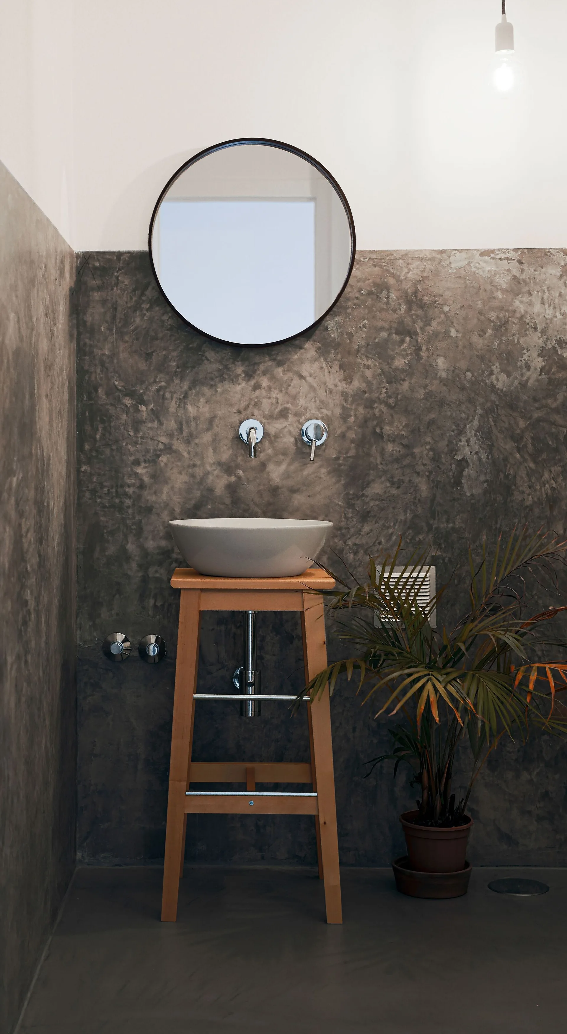 A white ceramic vessel sink rests on a tall wooden stool against a textured concrete wall in a minimalist industrial bathroom.