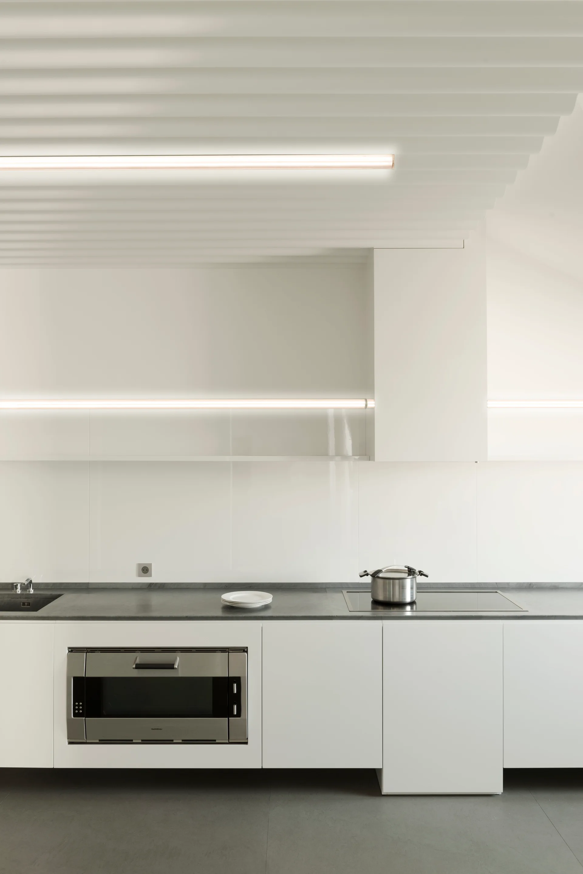 A minimalist white kitchen displays handleless cabinetry, a grey countertop, integrated appliances, and a slatted ceiling with linear strip lighting.