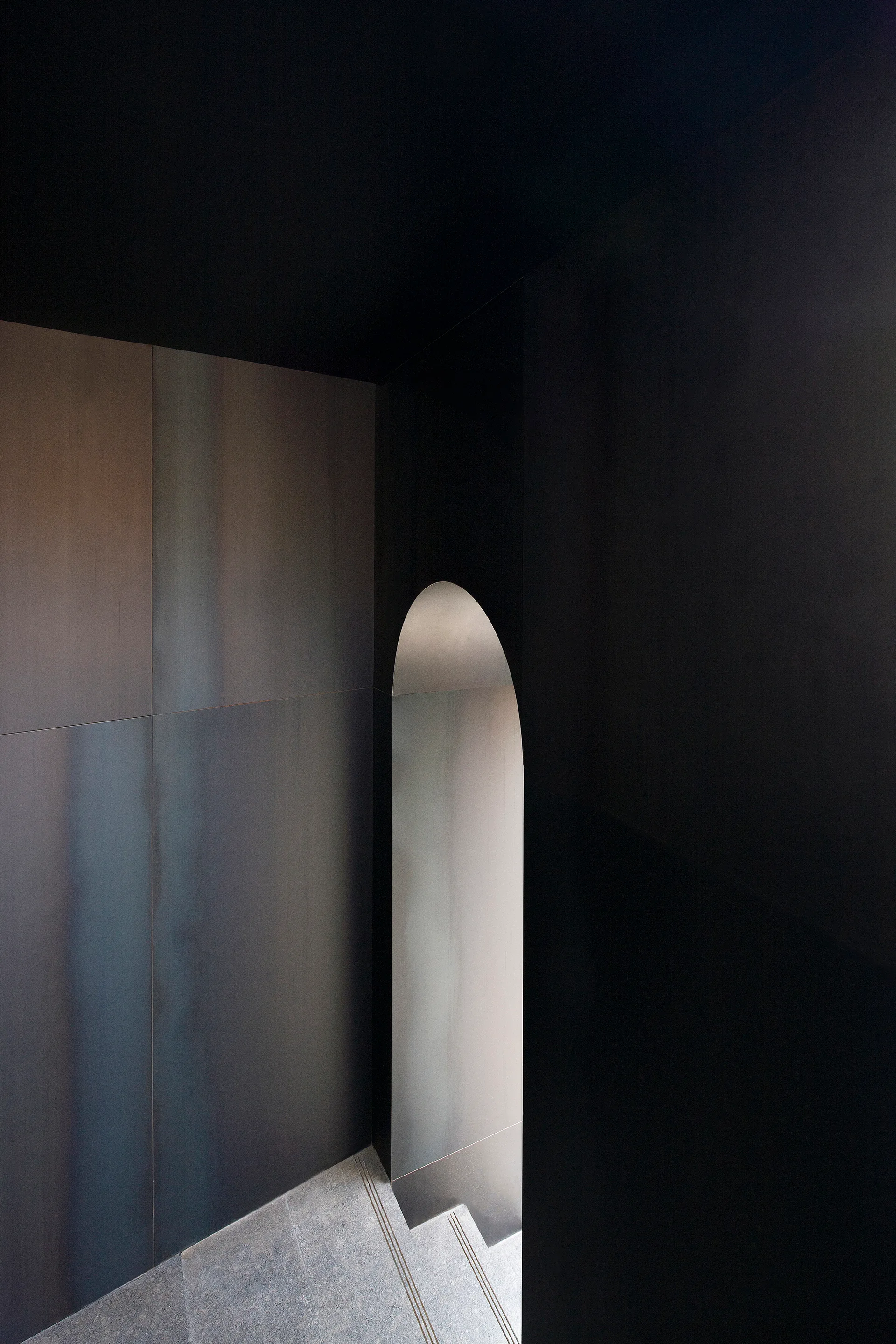 Dark grey wall panels line a minimalist staircase leading downward next to a smooth arched doorway.