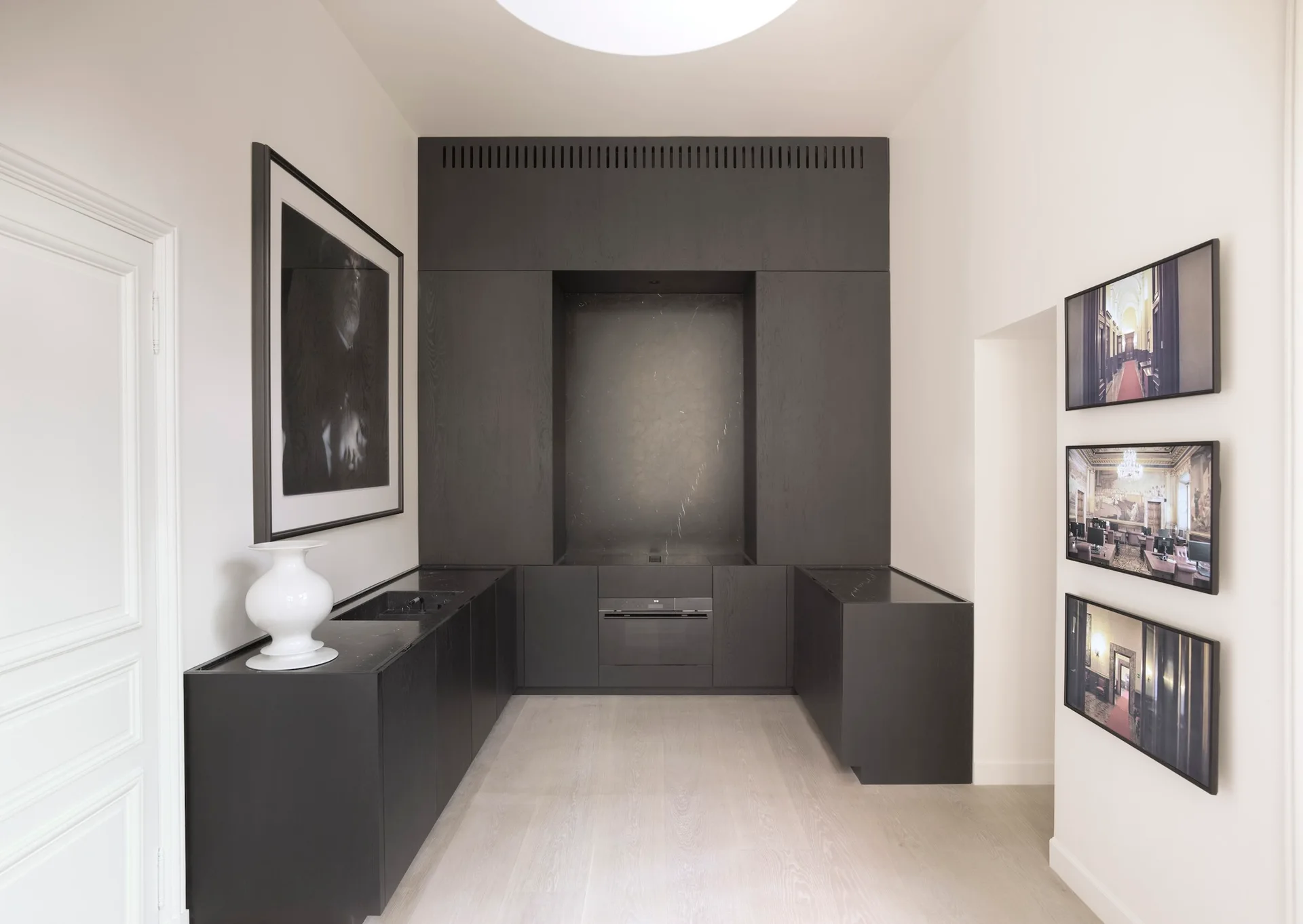 A minimalist kitchen features full-height matte black cabinets, integrated stone countertops, a central cooking niche, and light wood plank flooring.