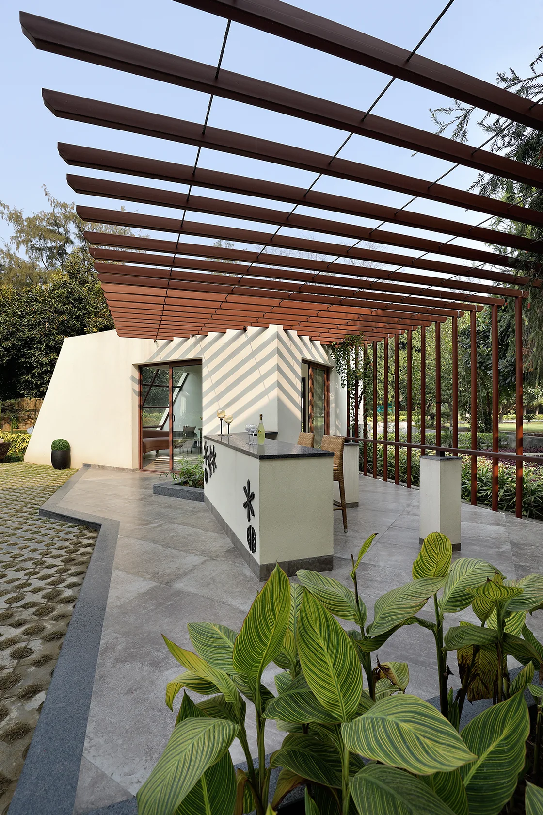 A contemporary outdoor patio bar with a beige counter and wicker stools sheltered by a brown slatted pergola casting linear shadows.