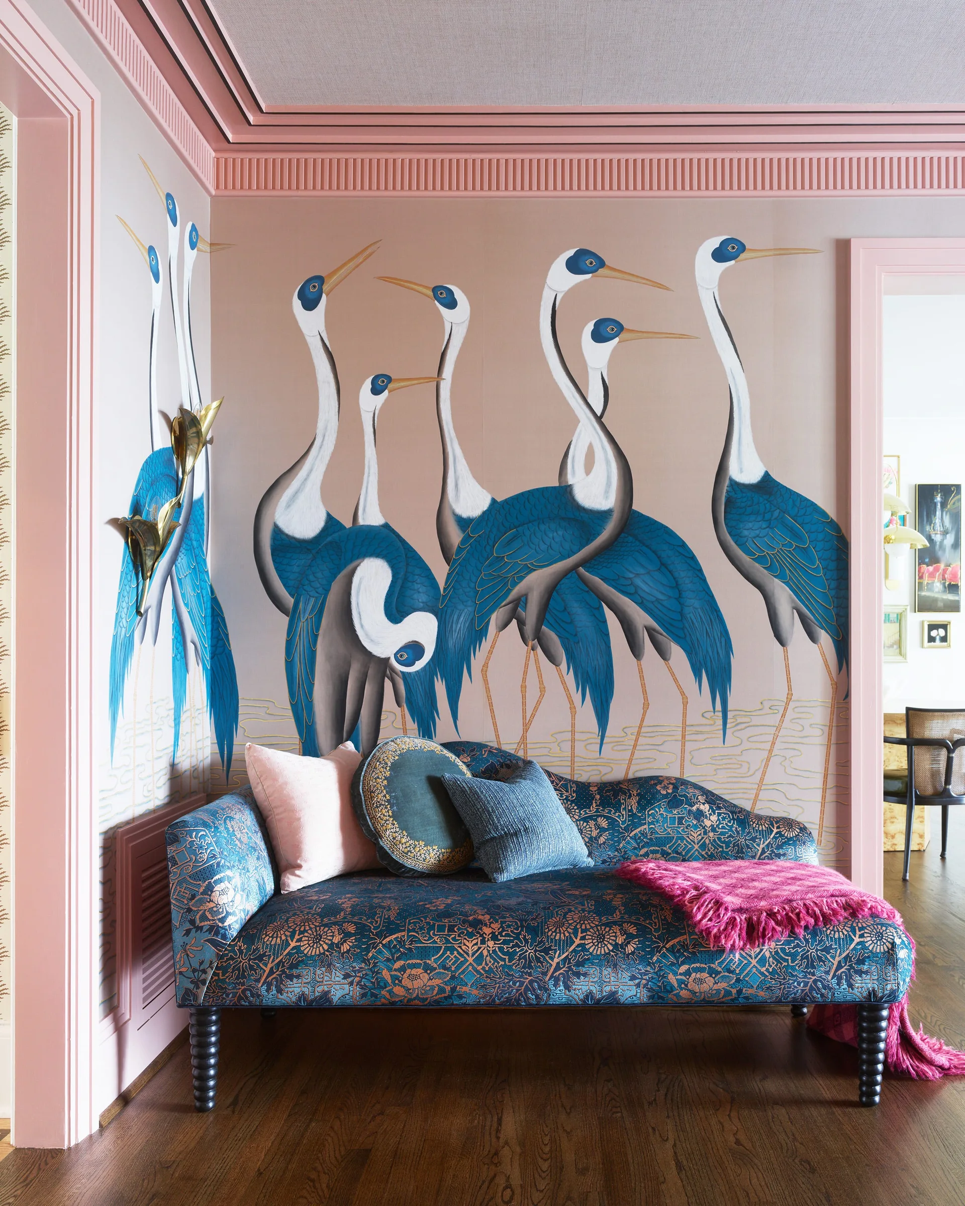 A blue patterned chaise lounge rests in a corner beneath a wall mural of blue and white herons framed by pink architectural molding.