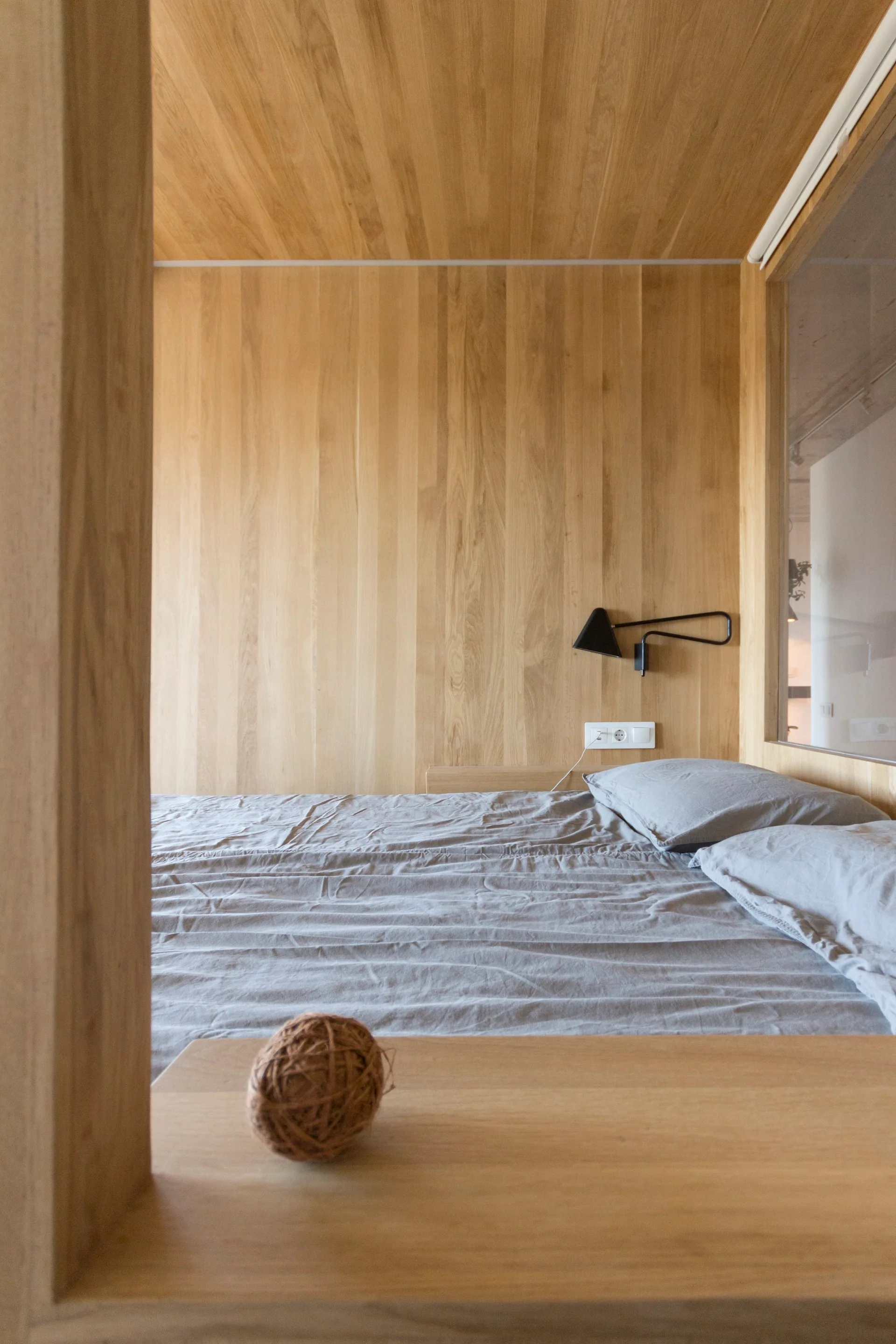 A minimalist bedroom nook features walls and ceiling clad in vertical light wood panels, grey bedding, and a black swing-arm wall lamp.