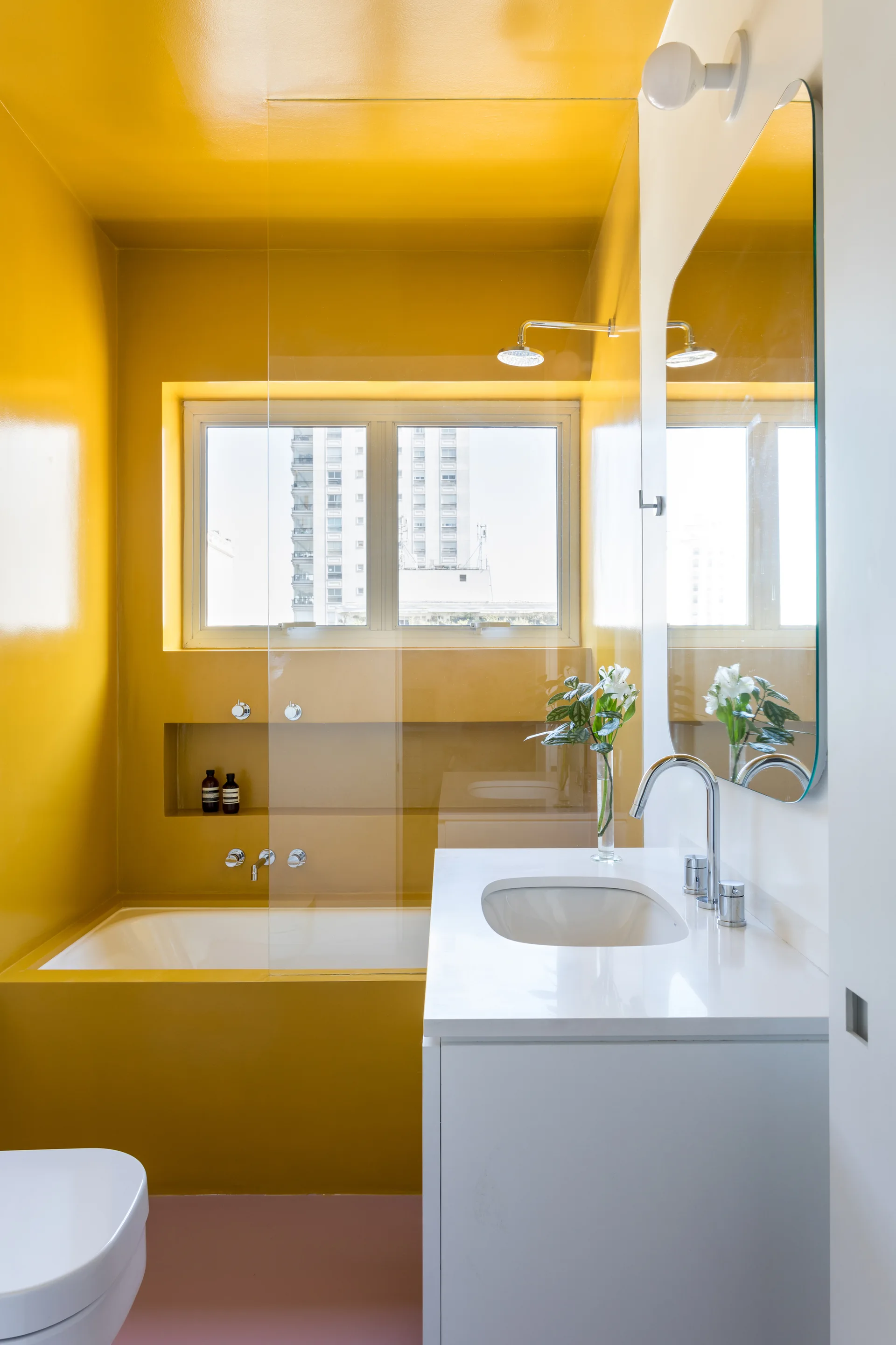A modern bathroom with glossy yellow walls, a white built-in bathtub under a window, a white vanity, and a frameless arched mirror.