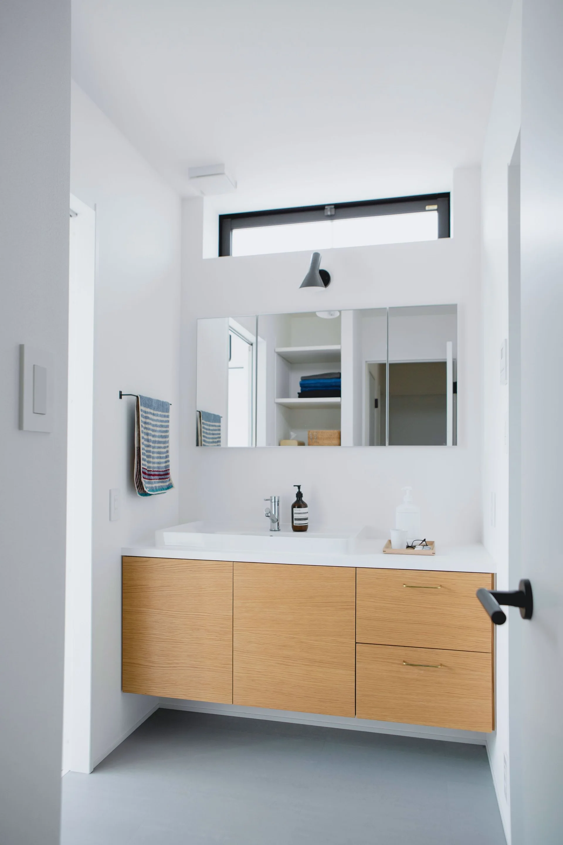 A minimalist bathroom with a floating light wood vanity, white integrated sink, mirrored cabinet, and a high horizontal window.