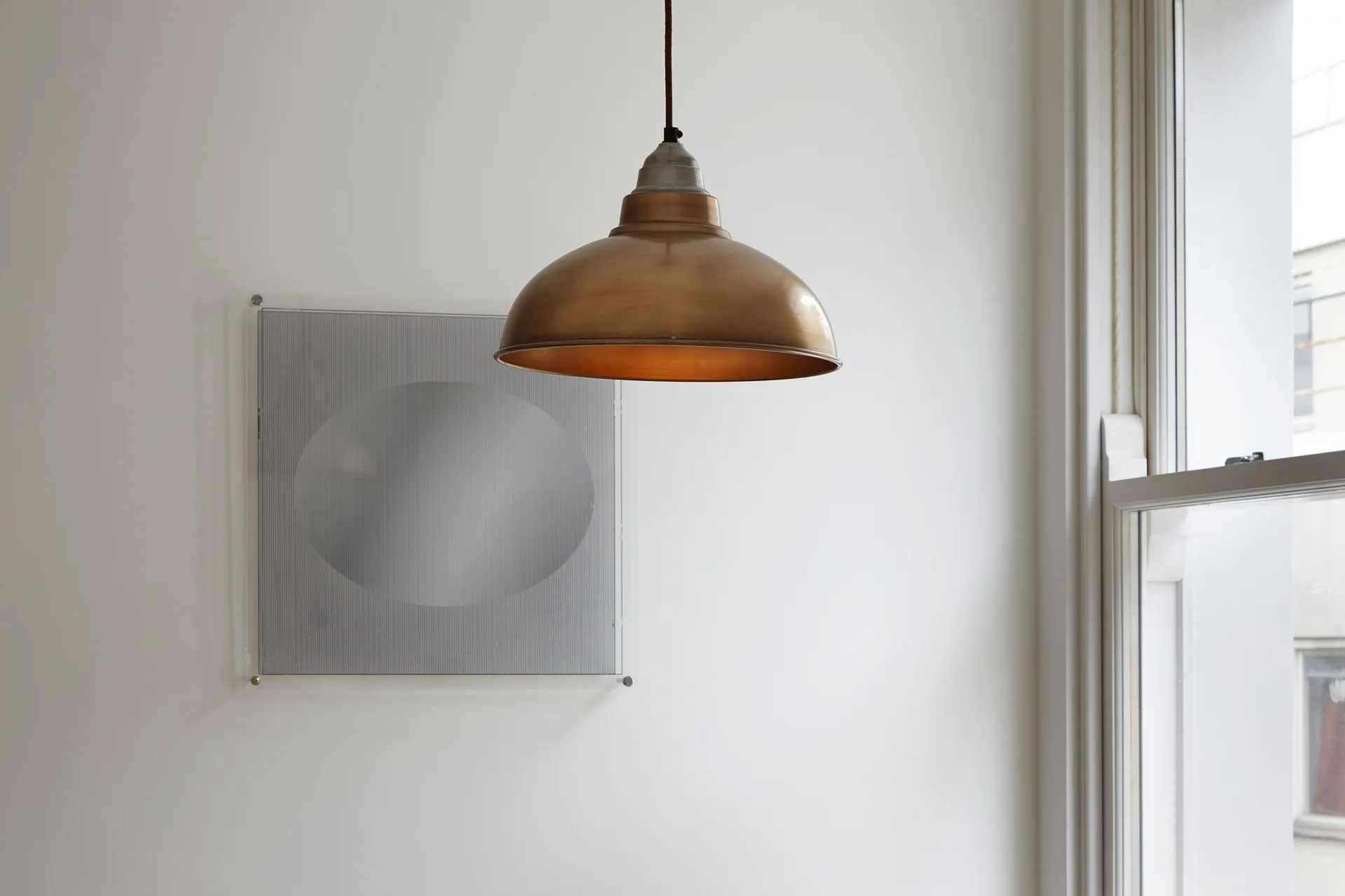 A metallic brass dome pendant light hangs in front of a white wall next to a window and a square piece of silver geometric art.