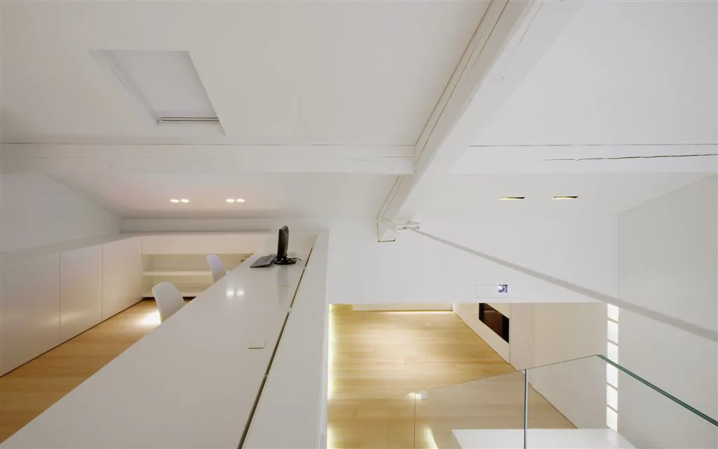 A high-angle view of a minimalist white loft home office featuring a built-in desk, light wood floors, and a glass railing overlooking the lower level.