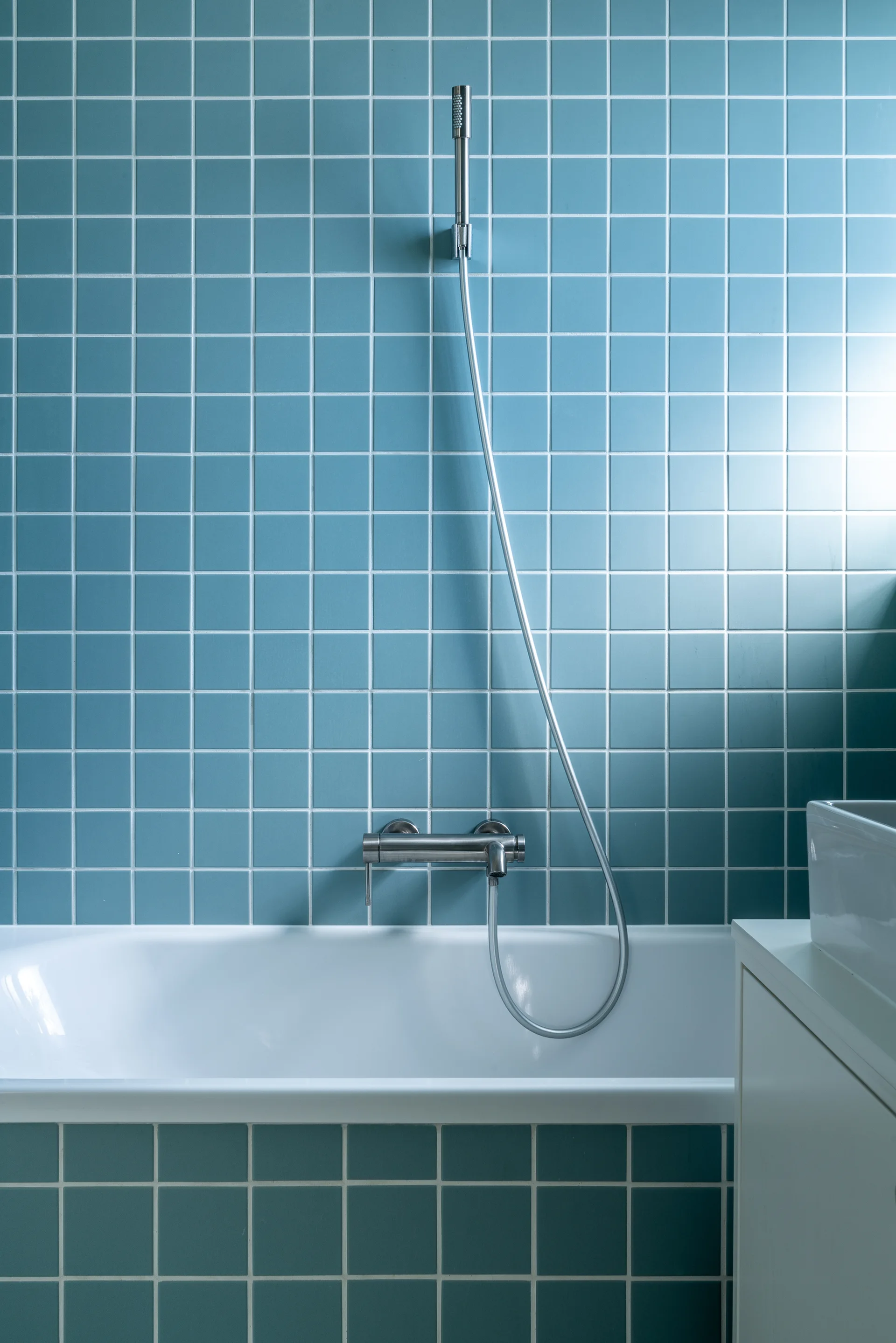 A close-up of a bathroom wall featuring blue square grid tiles, a wall-mounted stainless steel faucet, a handshower wand, and a white bathtub rim.