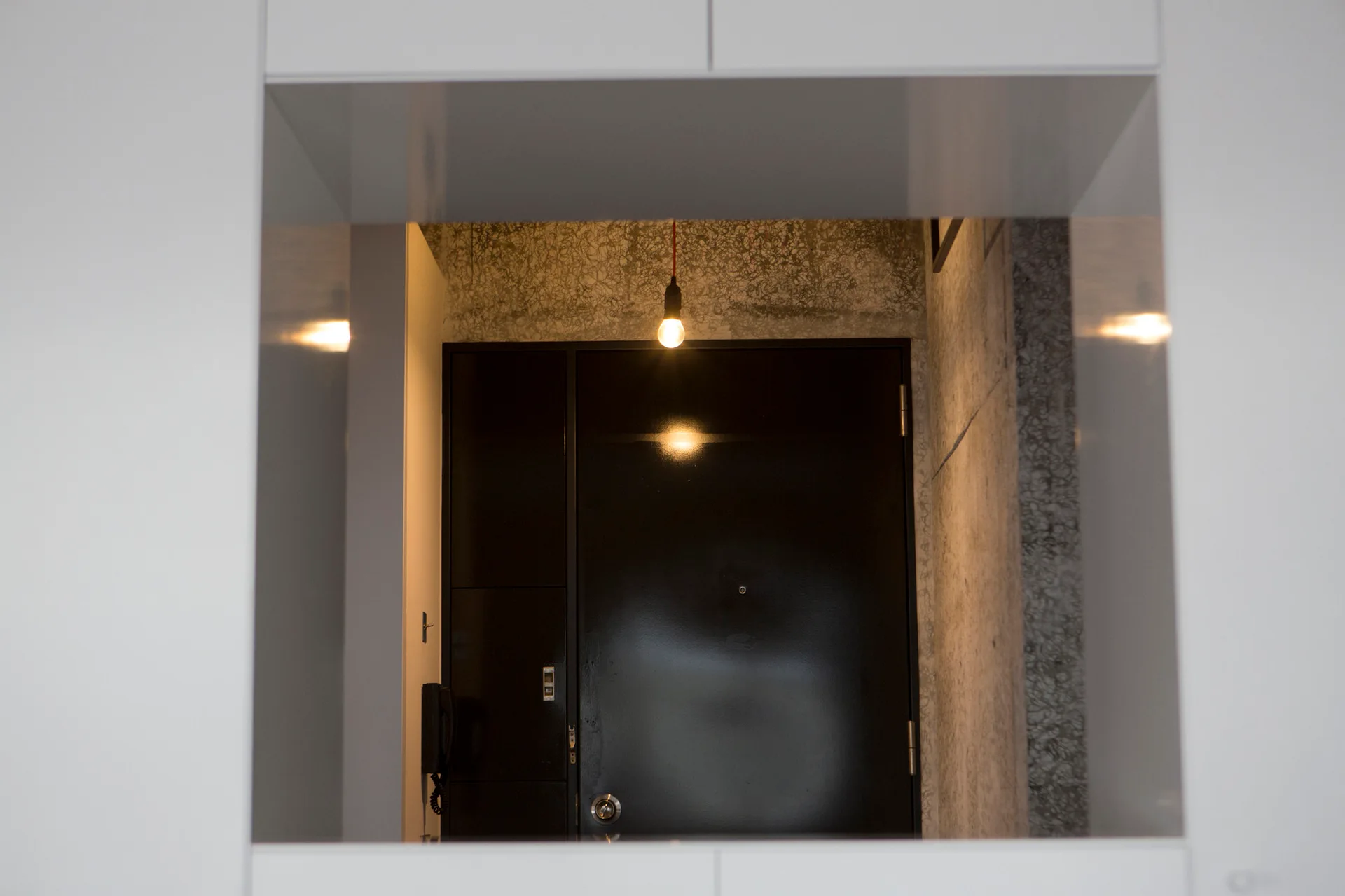 A view through a white rectangular frame shows an entryway featuring a black door, textured concrete walls, and a single hanging light bulb.