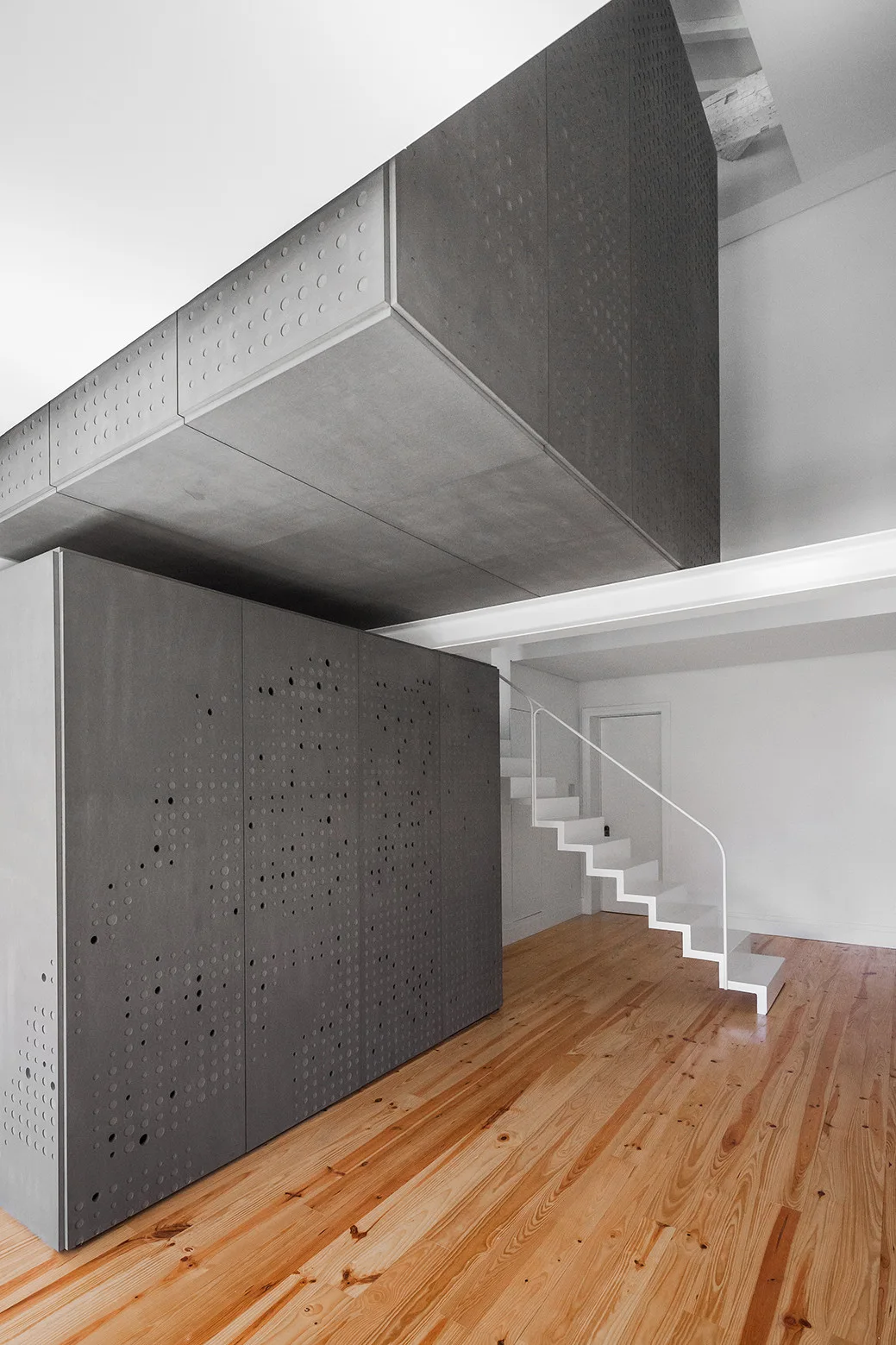 A minimalist white metal staircase ascends next to a large custom gray perforated storage cabinet on warm natural wood flooring.